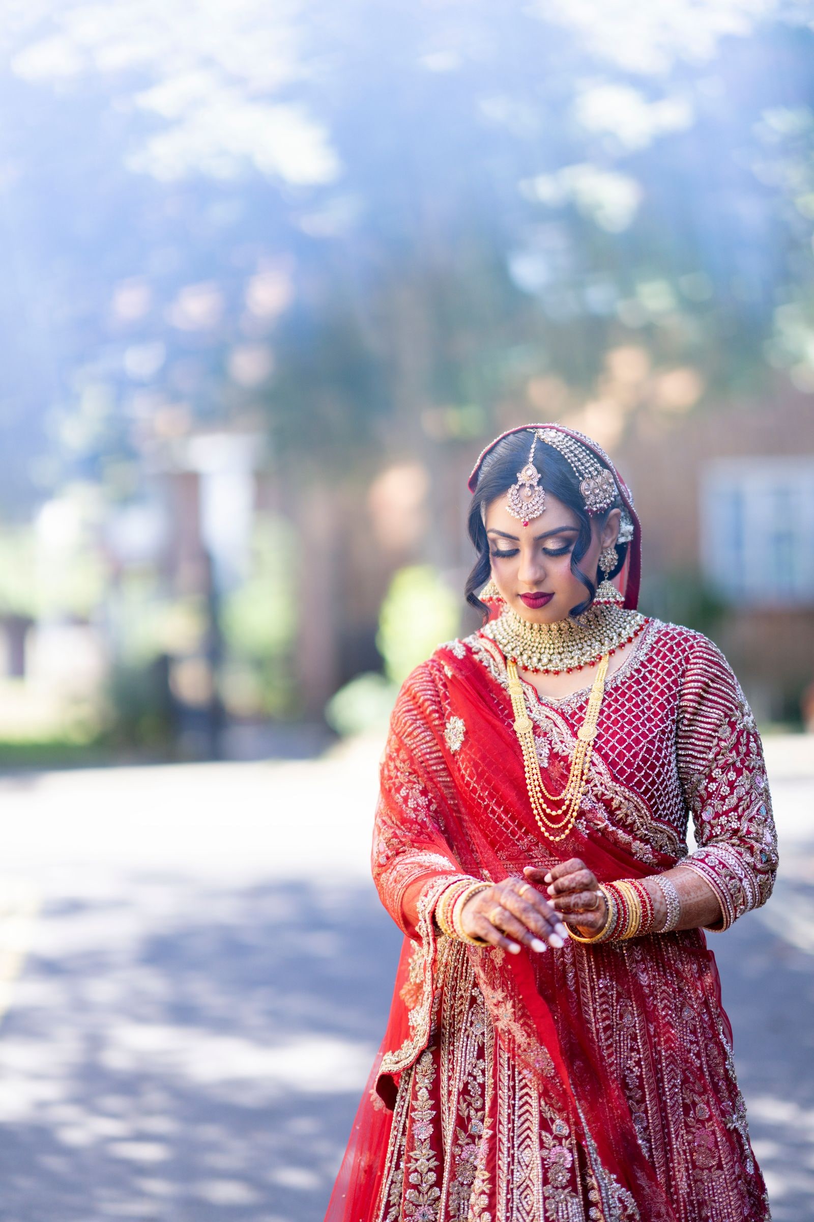 Bride in a red and gold lehenga on a sunlit residential street, eyes lowered
