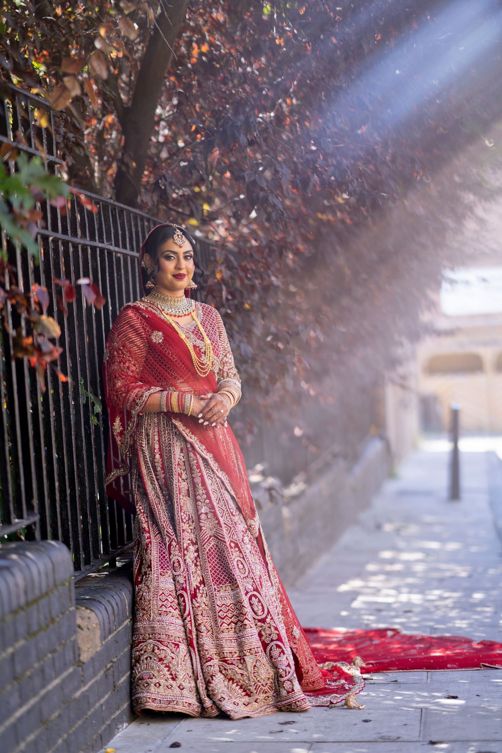 Bride leaning against a wall under a tree, her long red train spread on the pavement