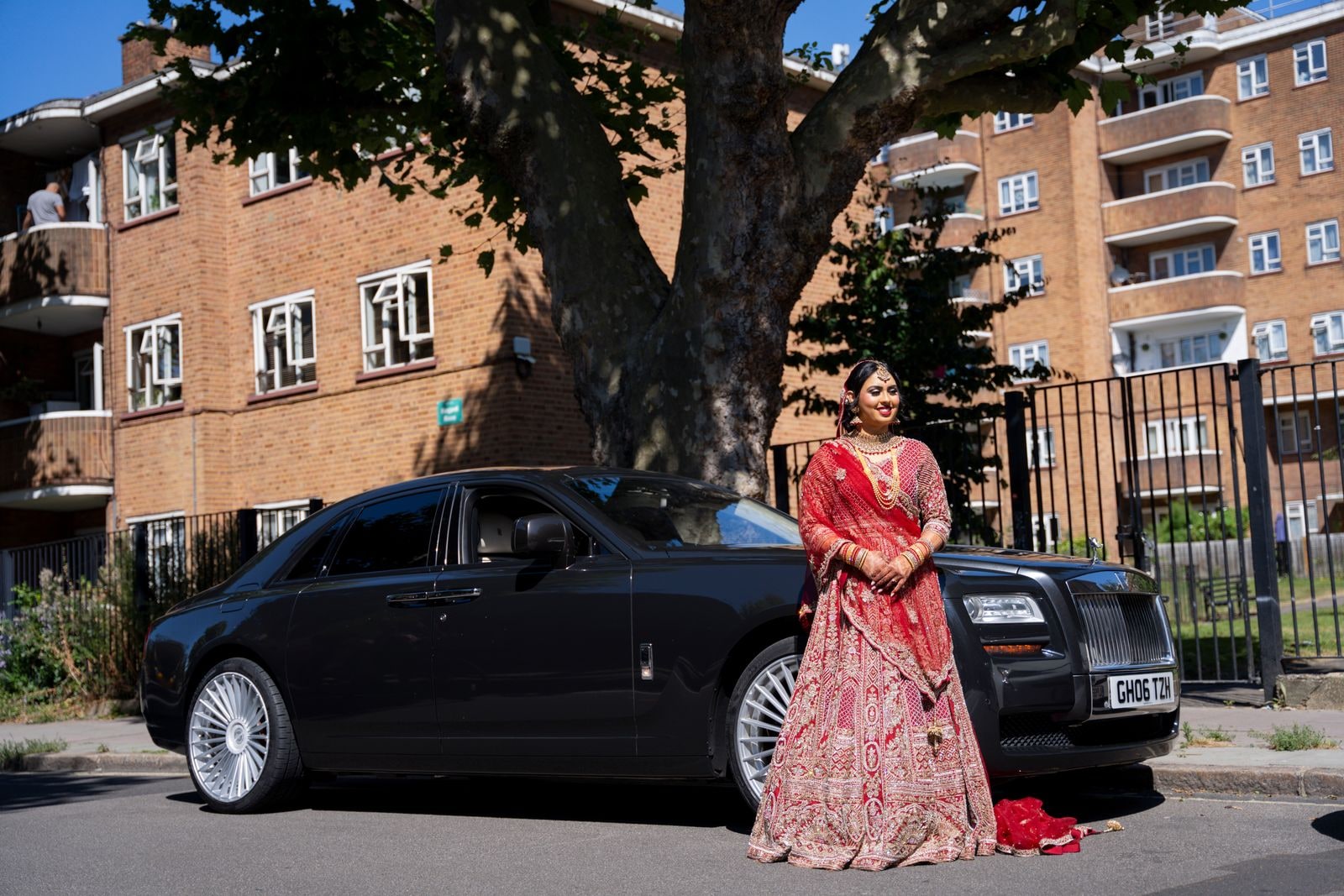 Bride in red beside a black Rolls-Royce parked under a plane tree