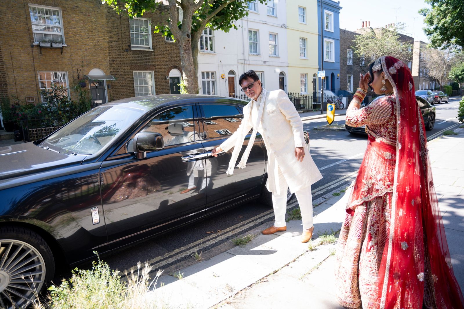 Groom in an ivory sherwani reaching for his bride across the front of the car