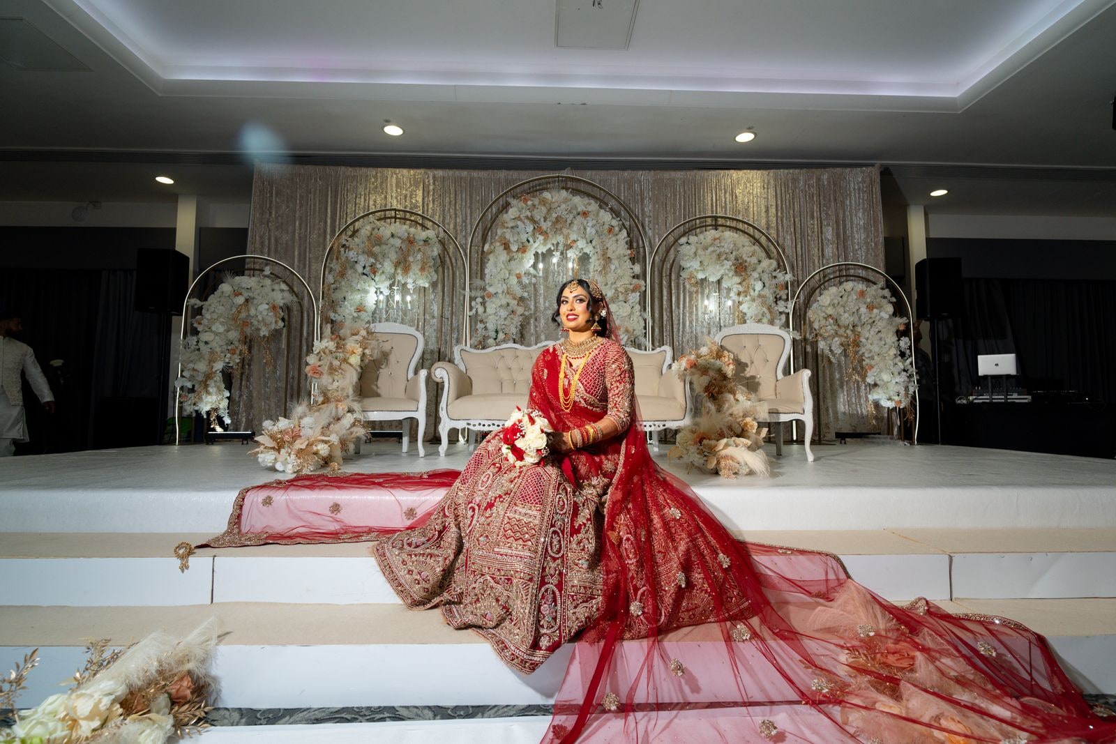 Bride seated alone on the flowered stage, her red train spilling down the steps
