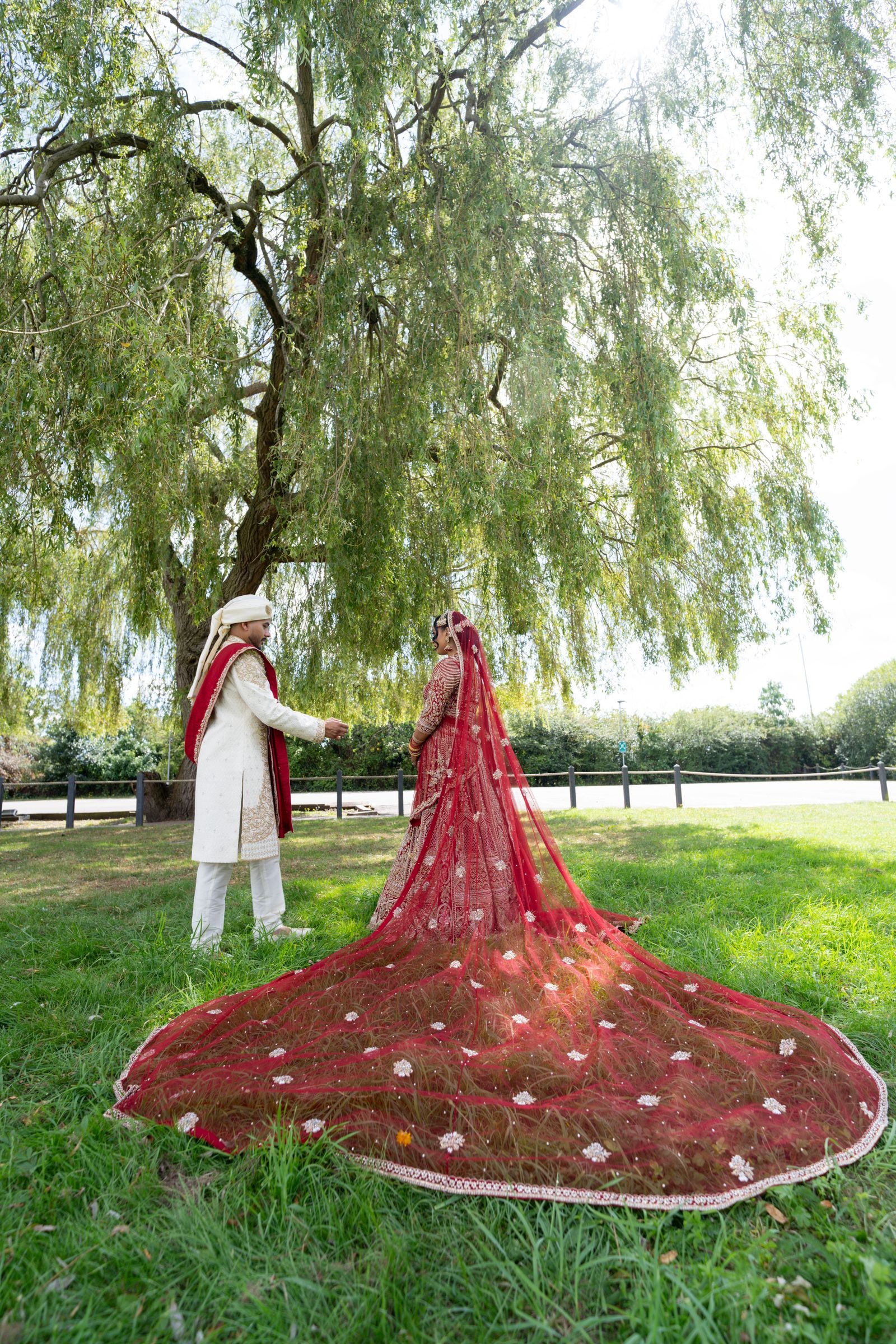 Groom holding his bride's hand under a willow, her red train laid out in a circle on the grass