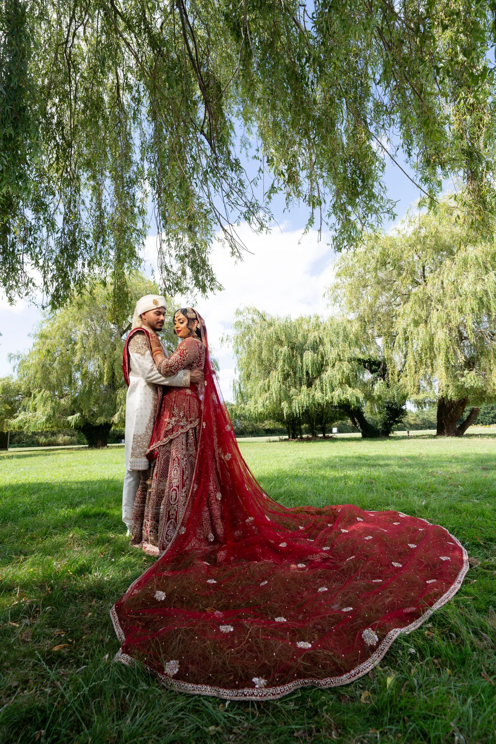Bride and groom holding each other under willows, her train spread wide behind her