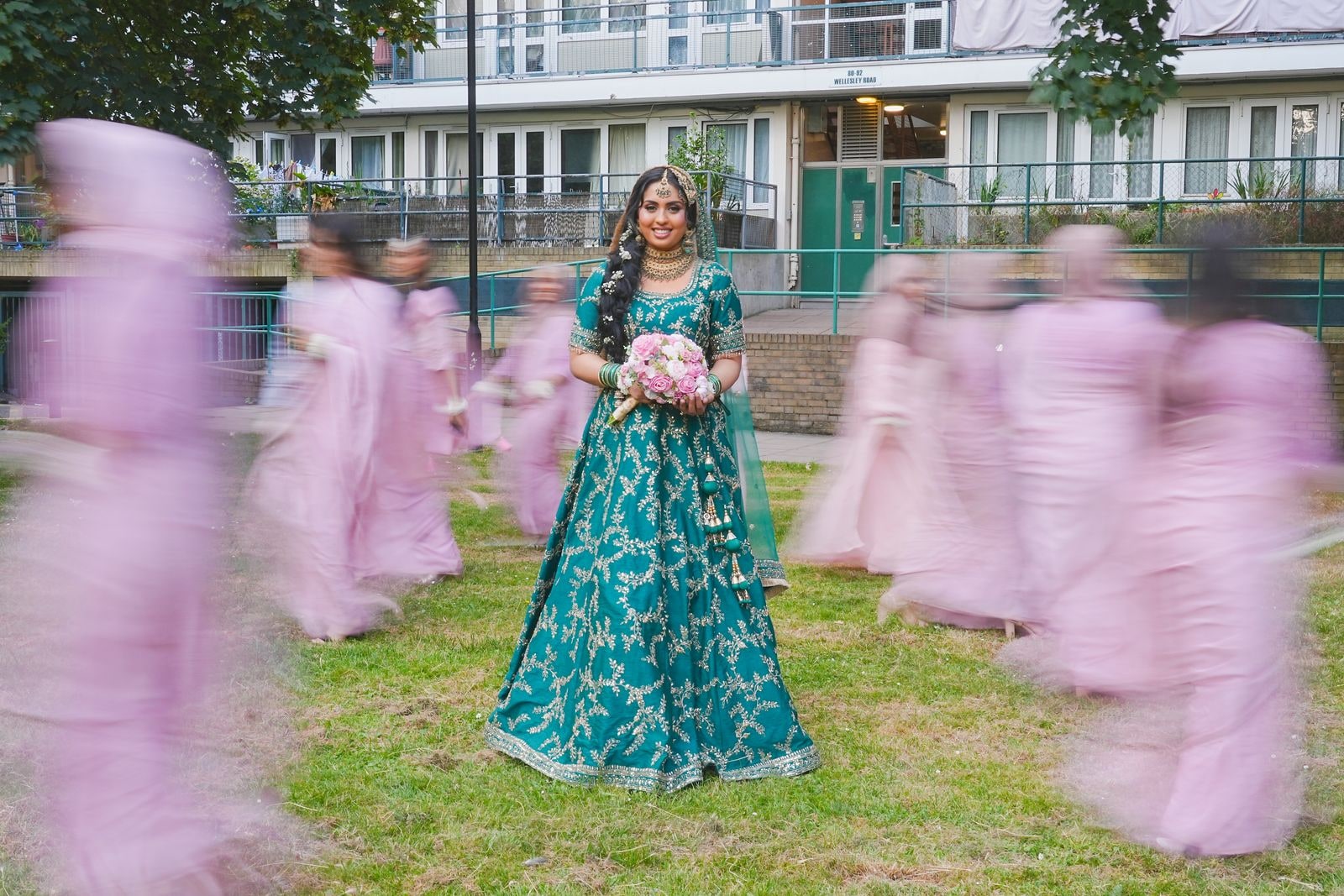 Bride in teal standing still while her party in pink circles her, blurred with movement