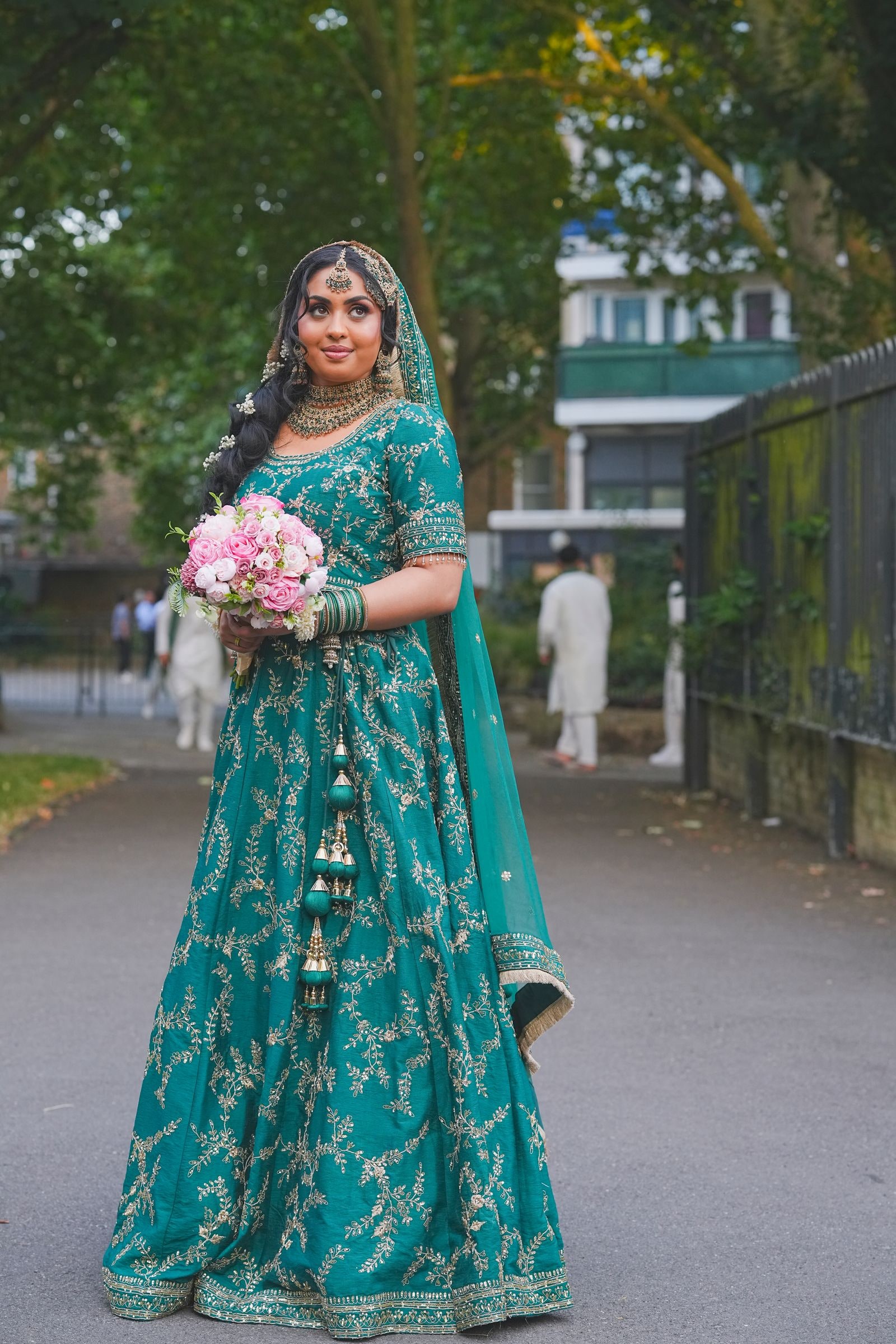 Bride in a teal and gold lehenga holding a pink bouquet, standing on a path