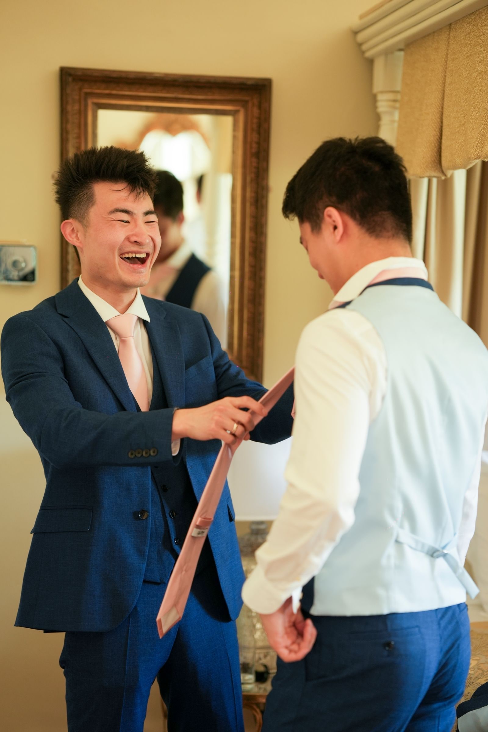 Groom laughing as a friend helps him with his tie in front of the mirror