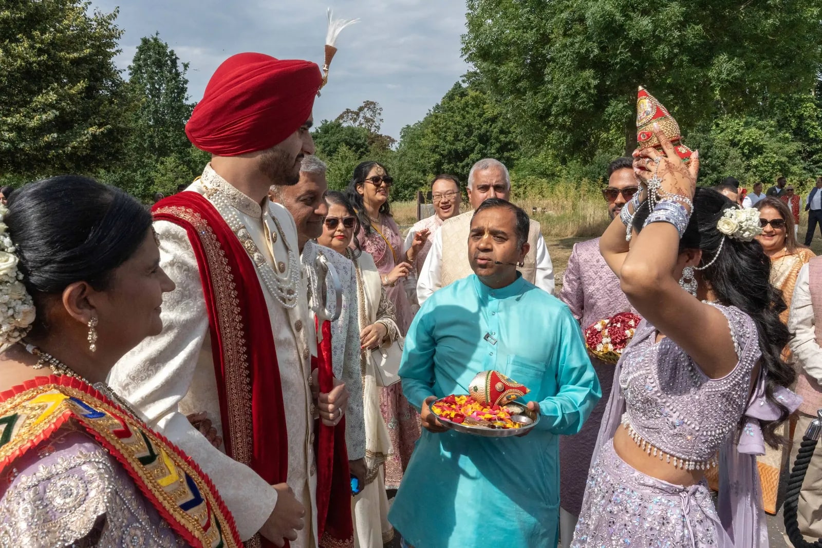 Groom in a red turban being welcomed with a tray as the family greets him