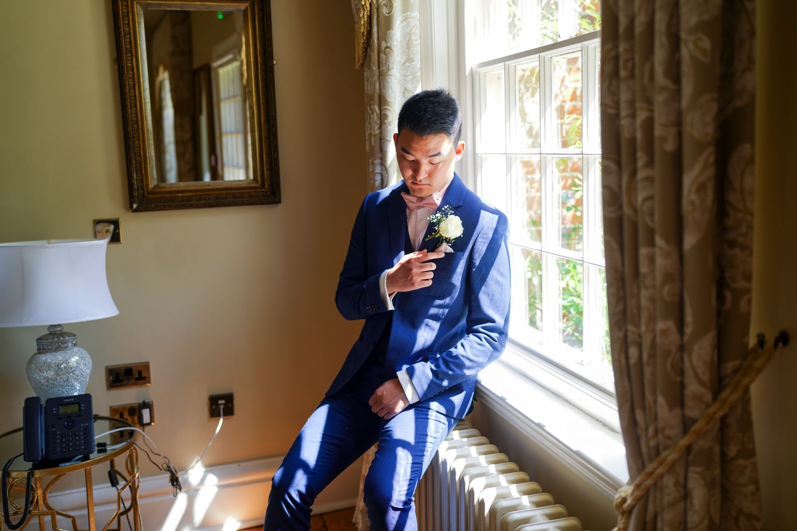 Groom in a blue suit fastening his buttonhole by a sunlit window