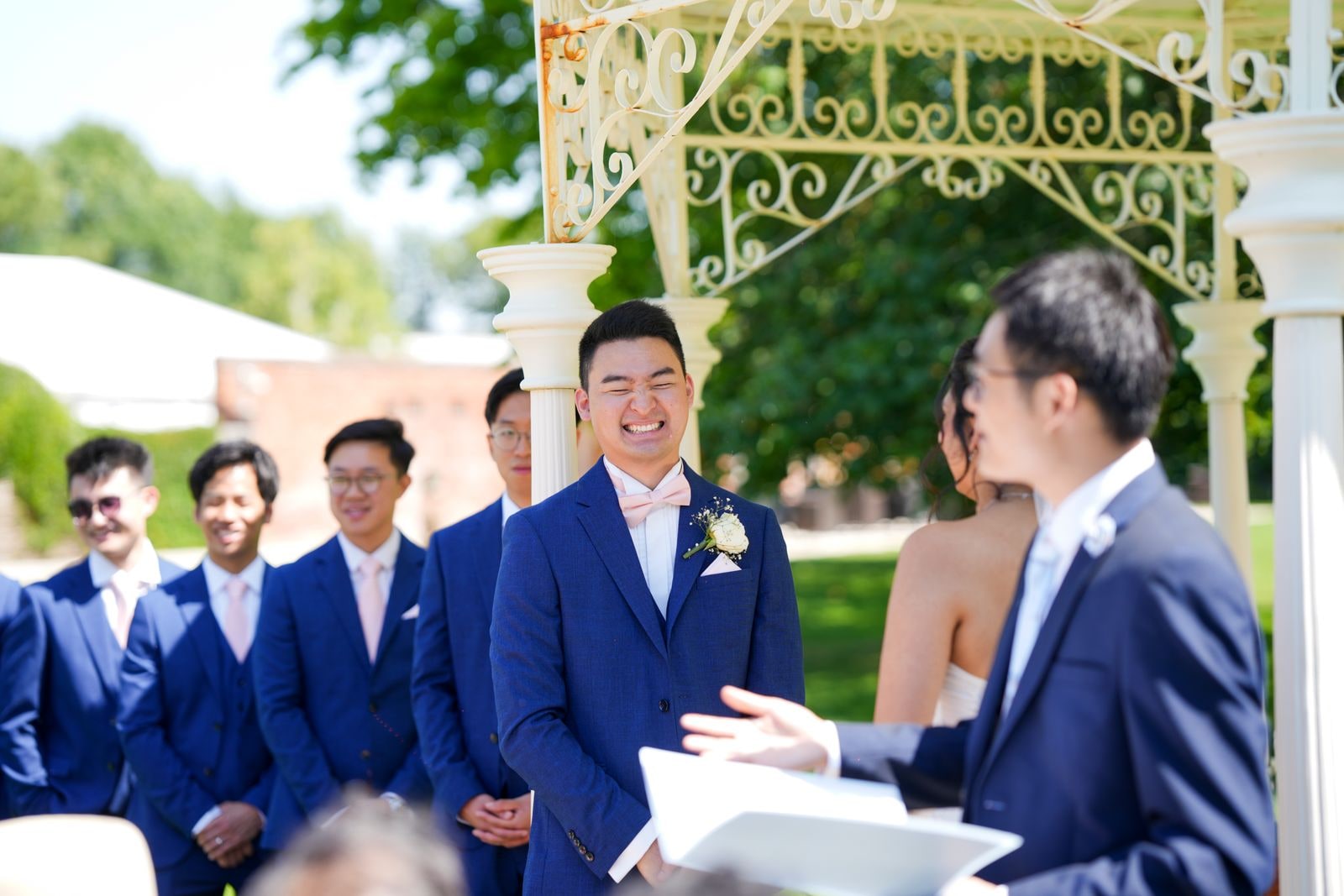 Groom smiling as the celebrant reads under the bandstand, groomsmen seated behind