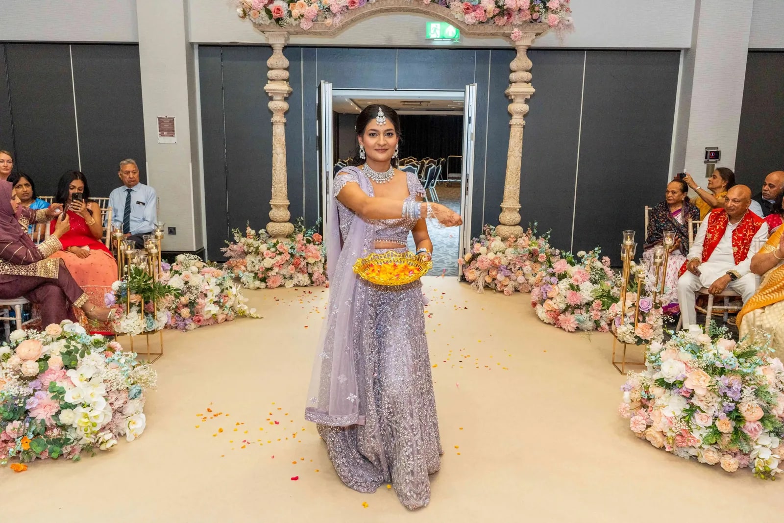 A woman in lilac walking the aisle carrying a bowl of marigolds