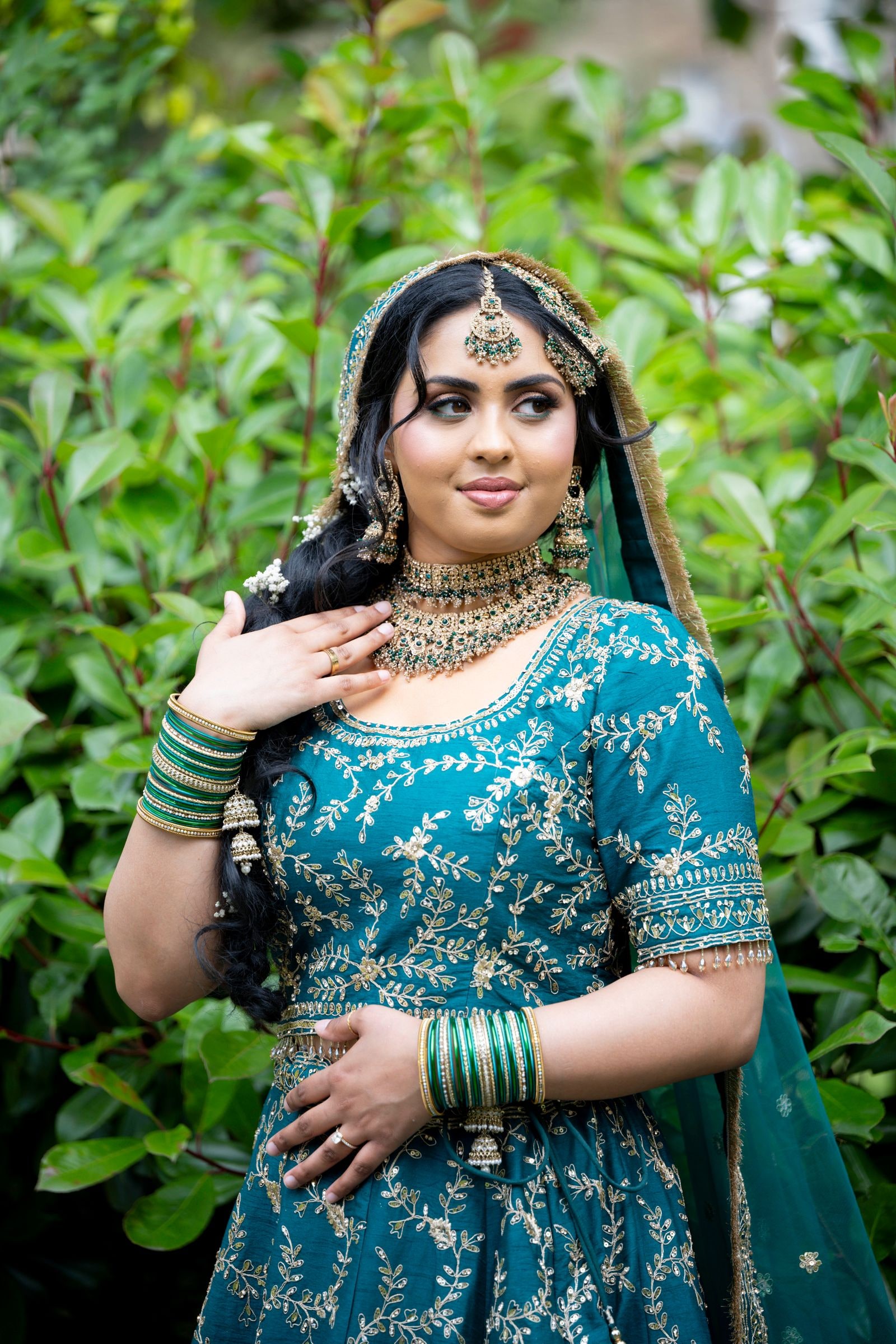 Bride in a teal and gold lehenga with green bangles, standing against leaves
