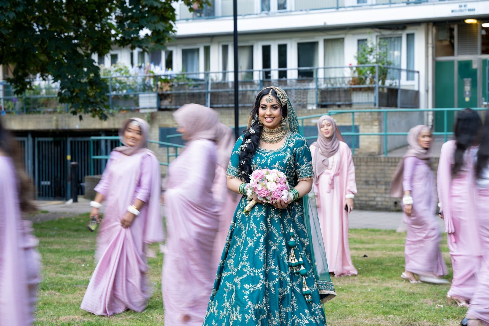 Bride in teal walking across the grass, her party in pink moving around her