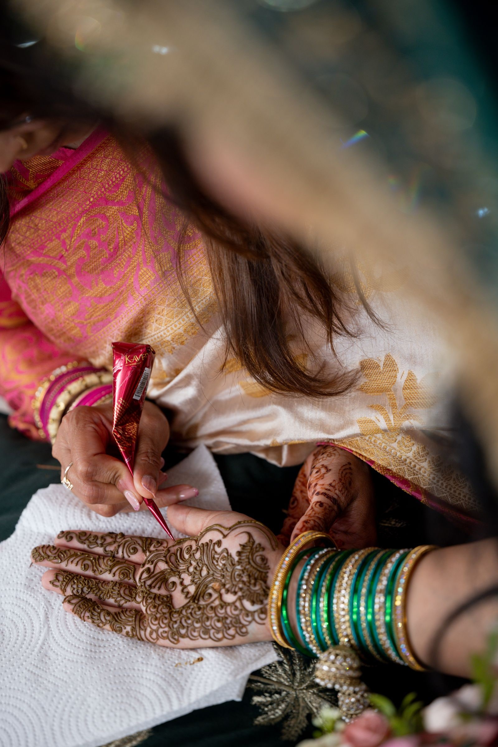 Henna being drawn on the bride's hand, her green bangles stacked at the wrist
