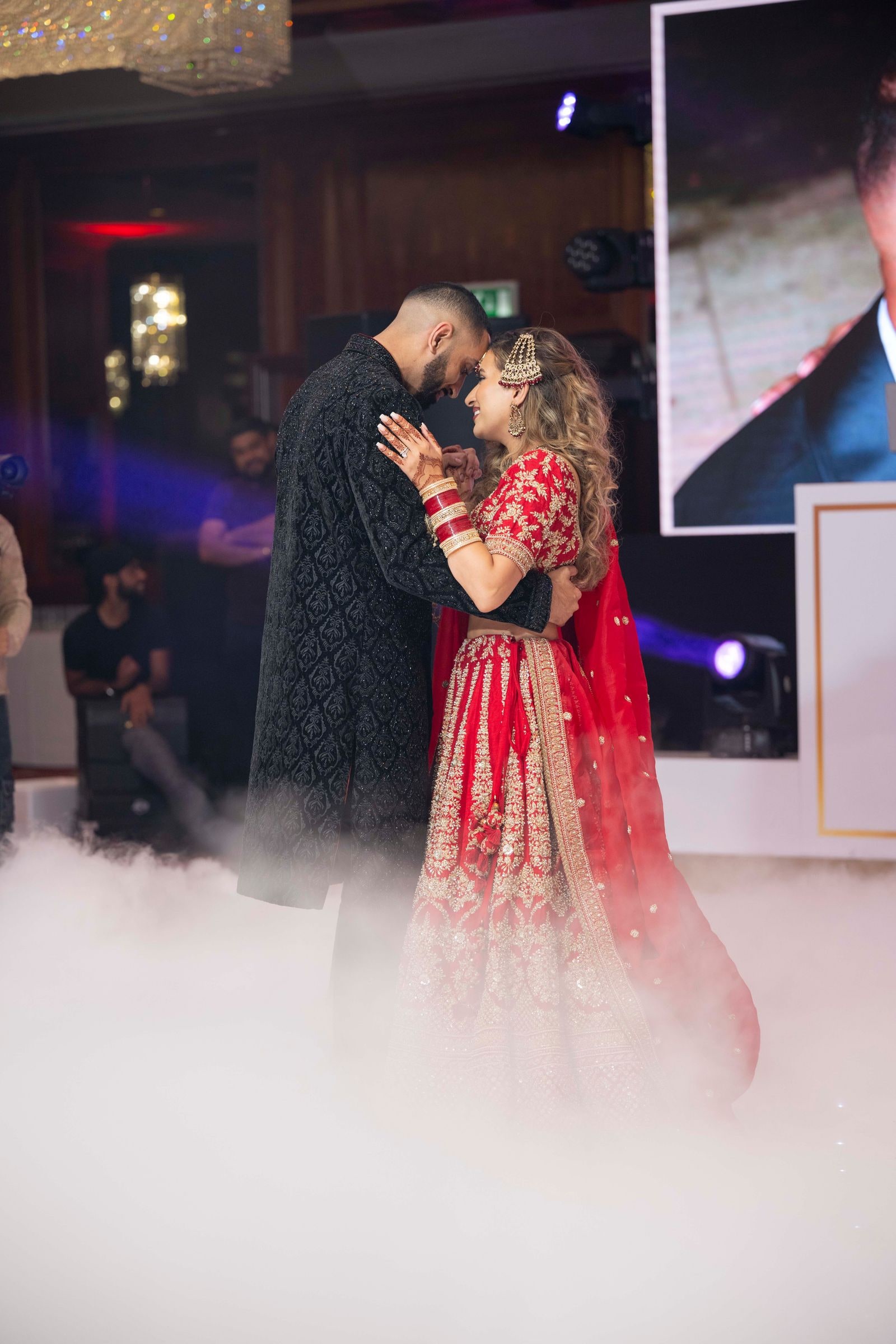 Couple dancing in dry ice, she in a red and gold lehenga, he in a black sherwani
