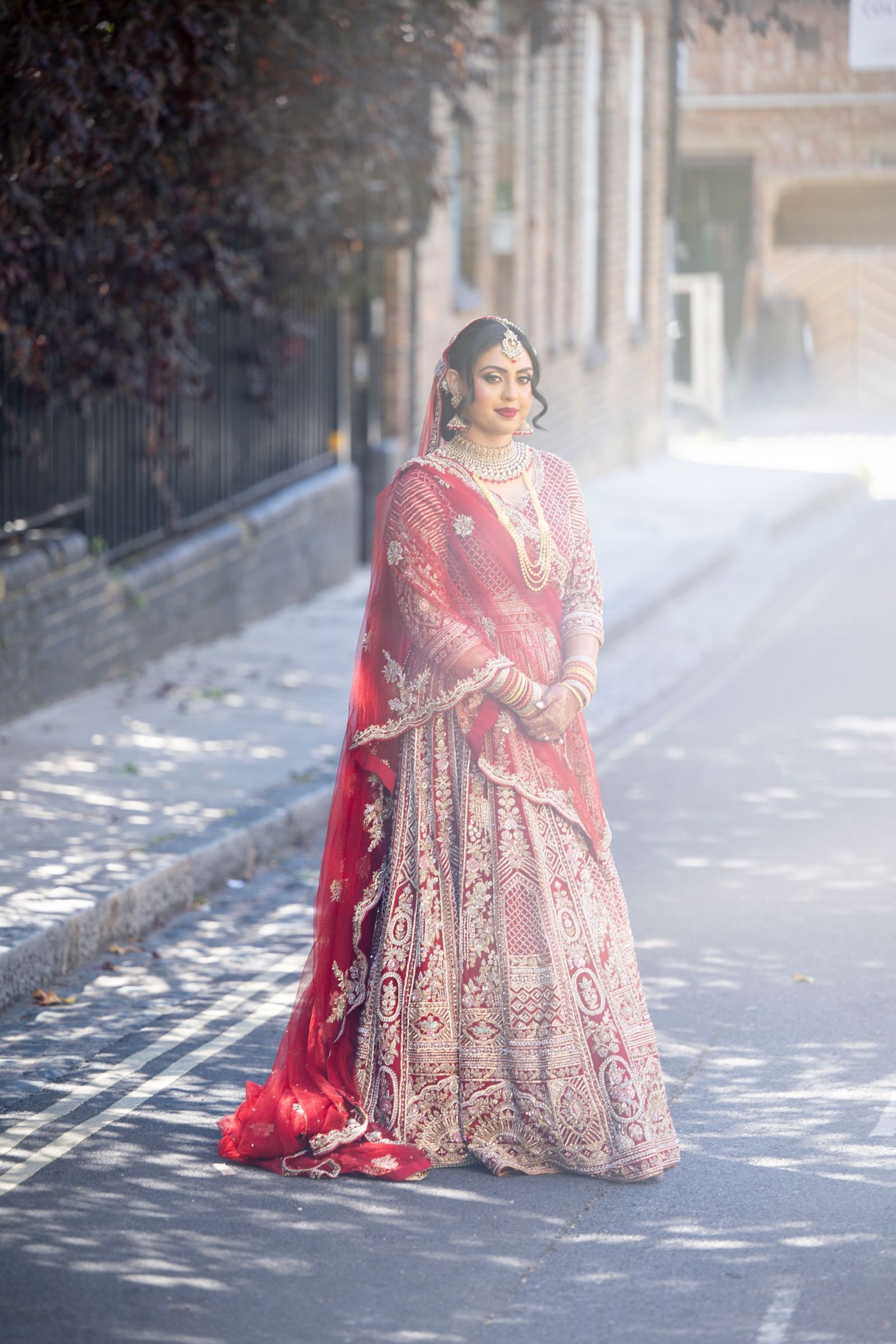 Bride in a blush and red lehenga standing in an empty street in the morning light