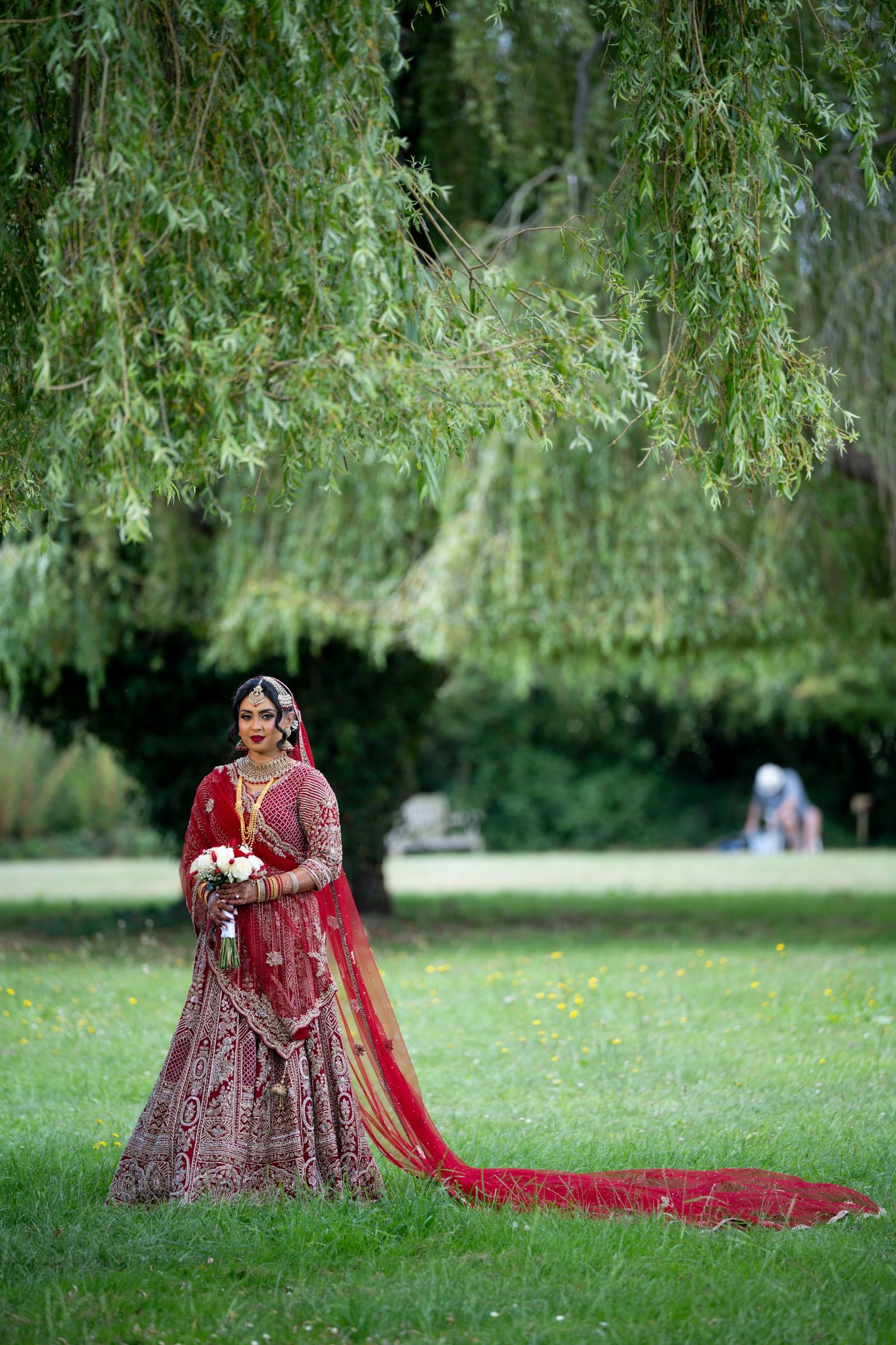 Bride in deep red beneath a willow, her long train laid out across the grass