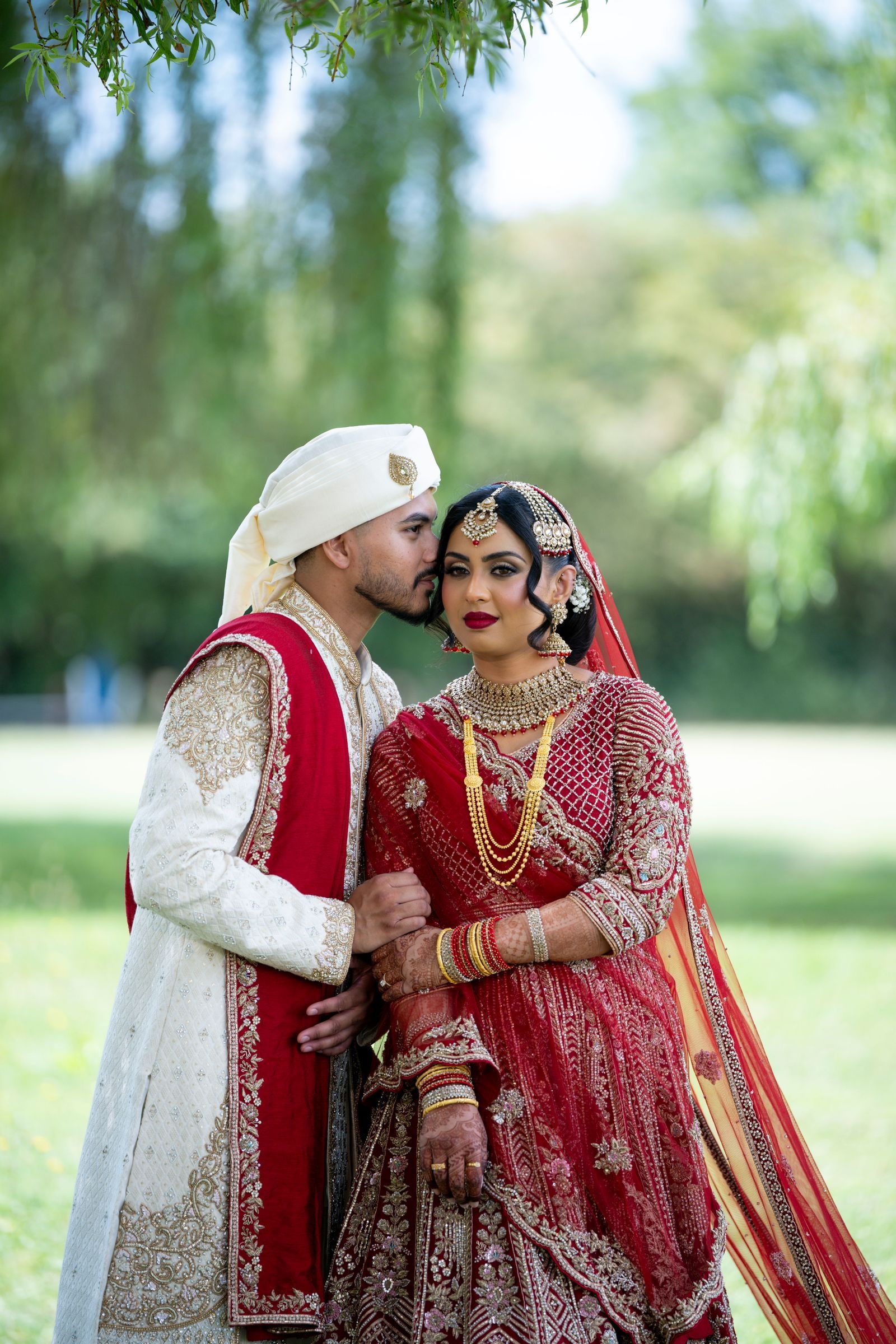 Groom kissing his bride's cheek under a willow, both in red and ivory