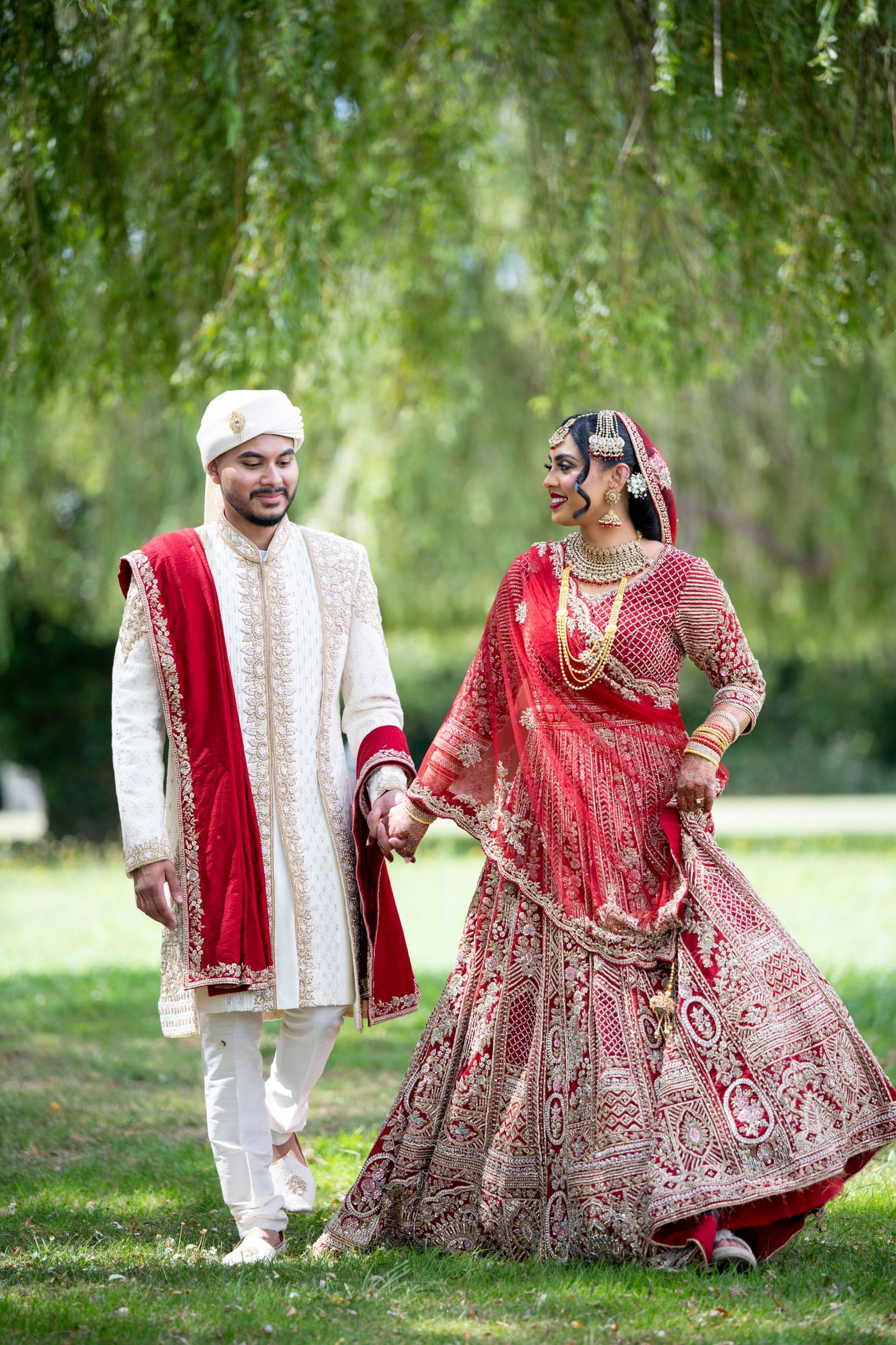 Bride and groom walking hand in hand across the park in red and ivory