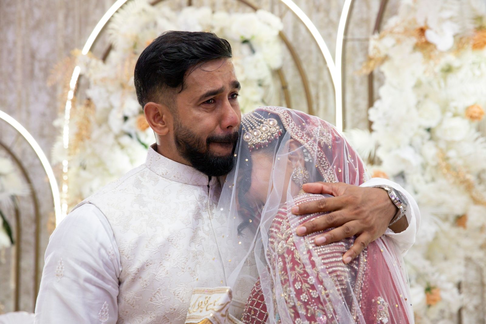 Groom in an ivory kurta wiping his eyes, his bride's veil against his shoulder