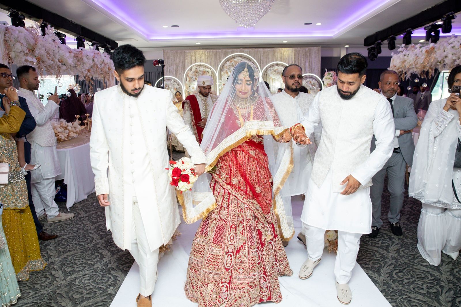 Bride walked down the aisle beneath her veil by two men in ivory sherwanis