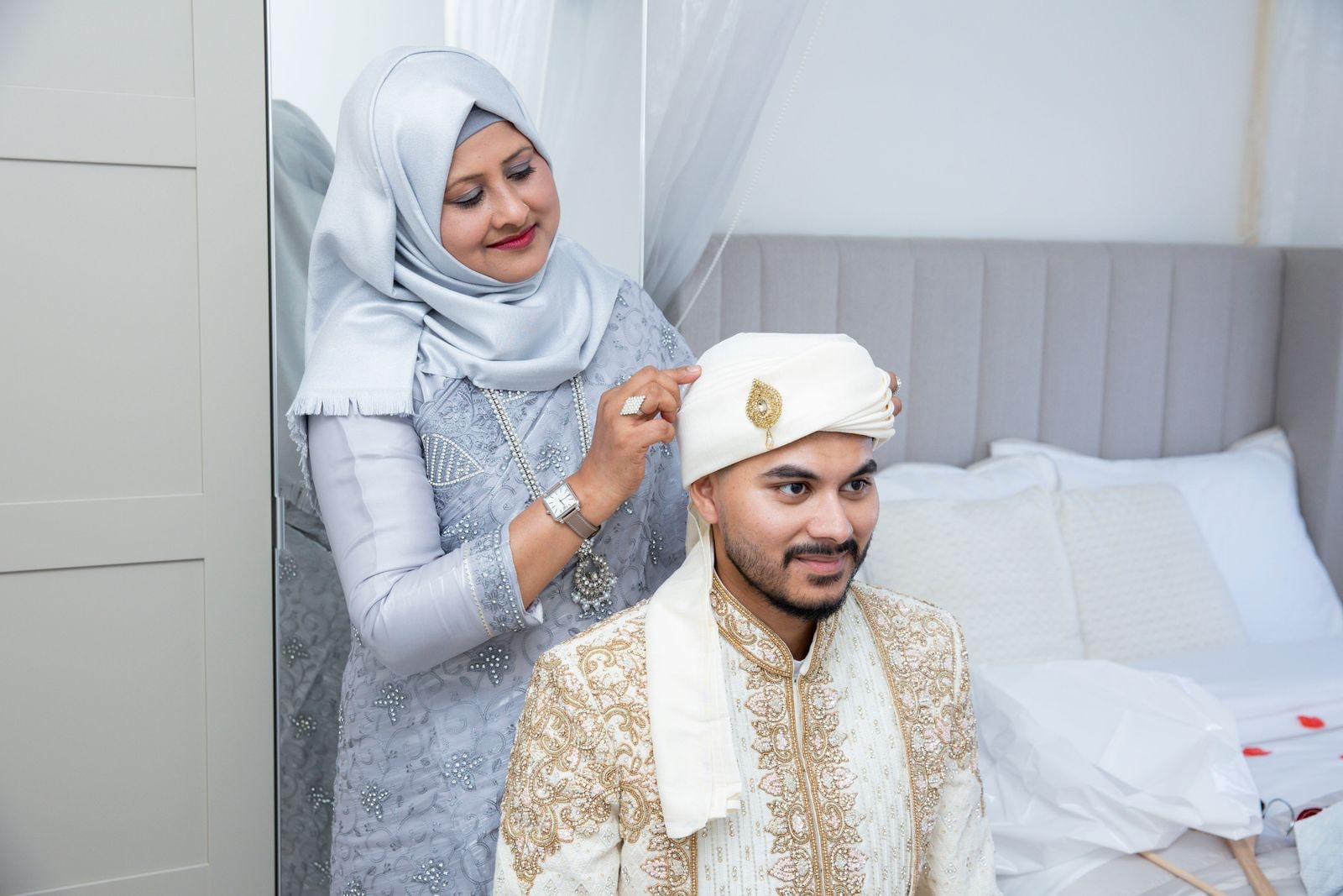 The groom's mother adjusting his topi as he sits on the bed