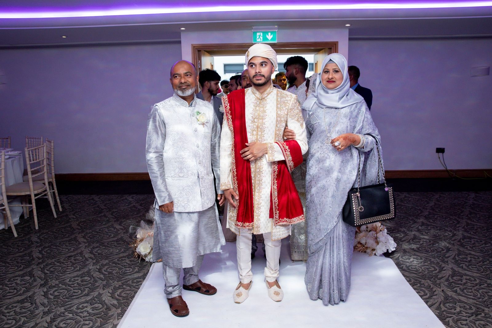 Groom walked in by his parents along the white aisle, in a red and ivory sherwani