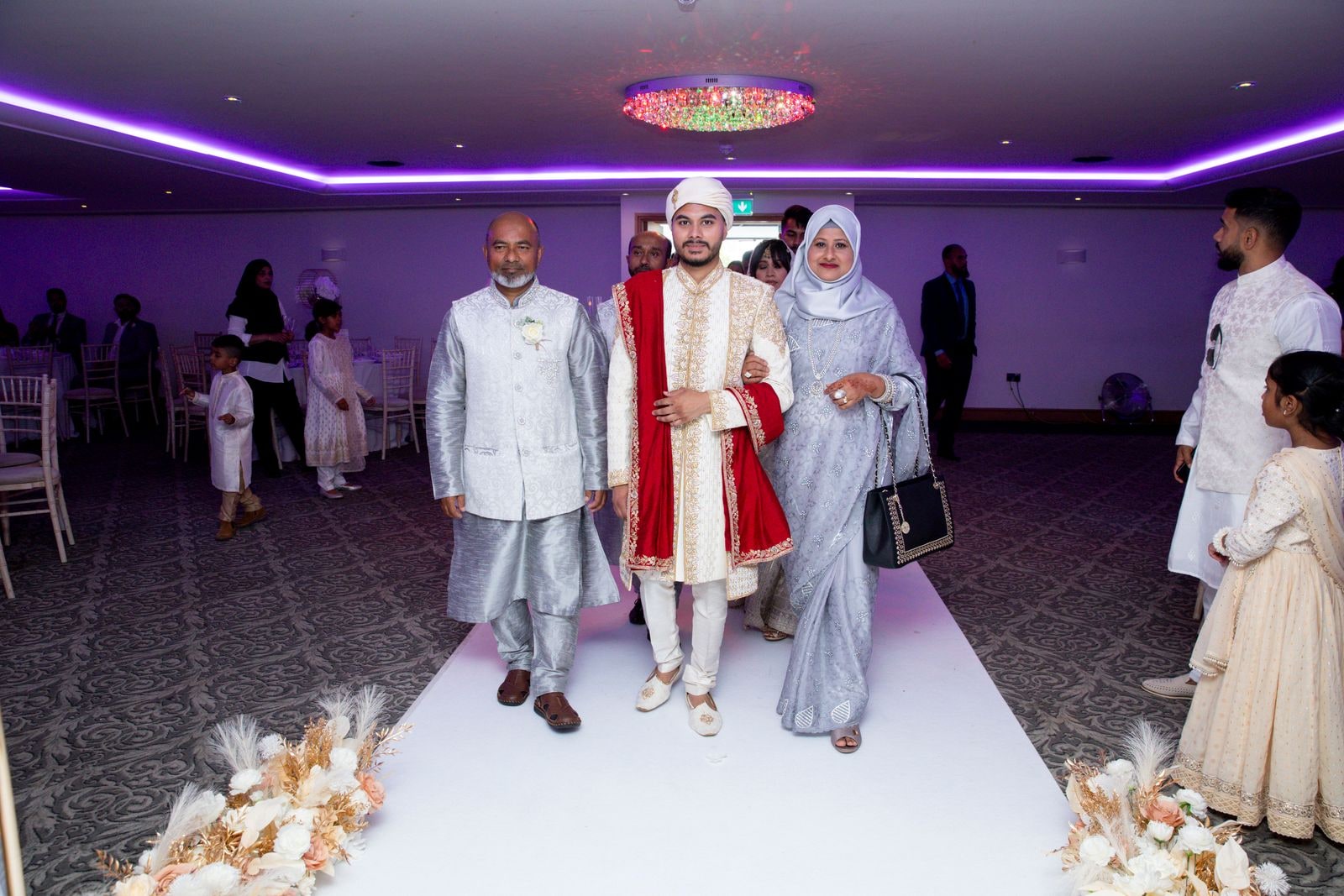 The groom's parents walking him into the hall as guests watch