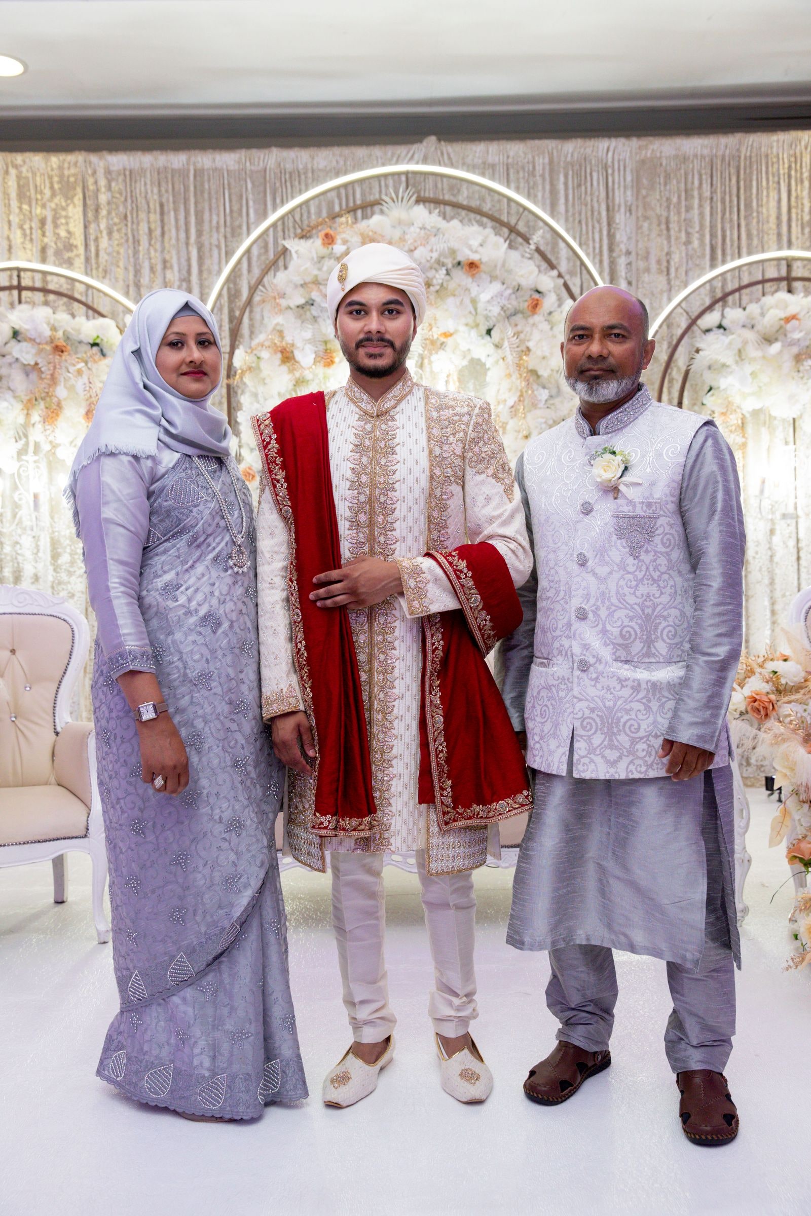 Groom standing between his parents in front of the flowered stage