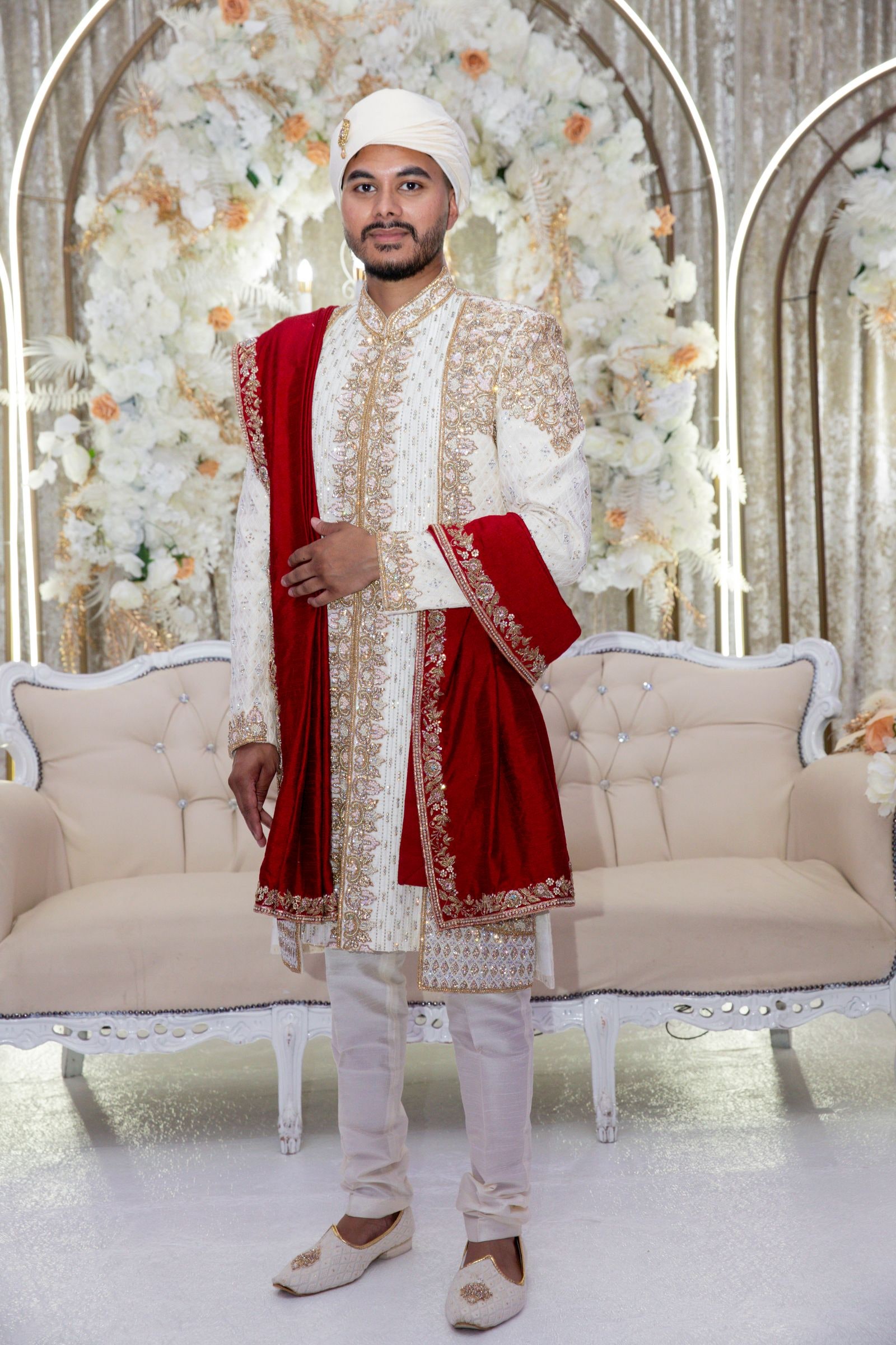 Groom alone before the white flower arch in an ivory sherwani and red shawl