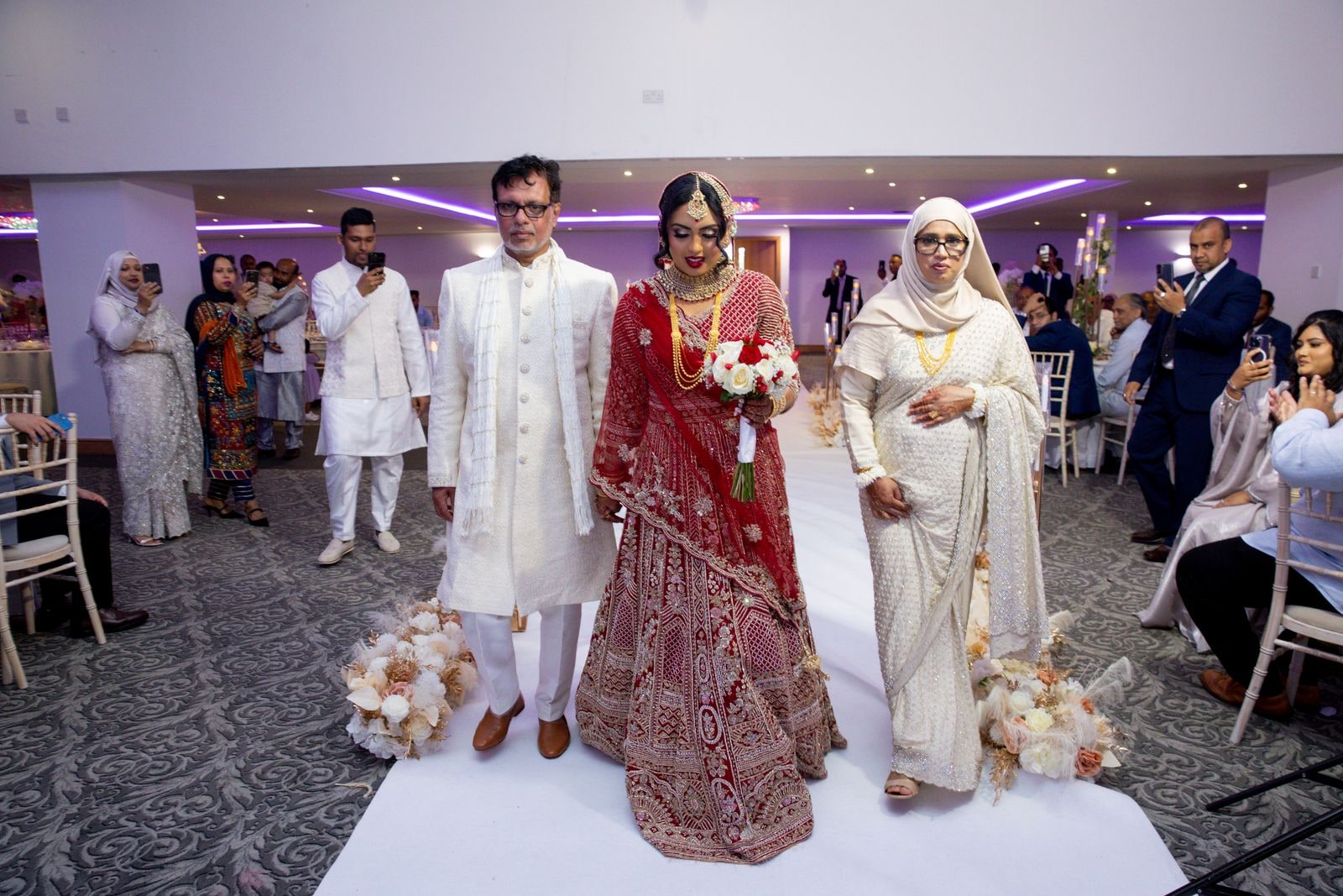 Bride between her mother and father on the white aisle, holding her bouquet