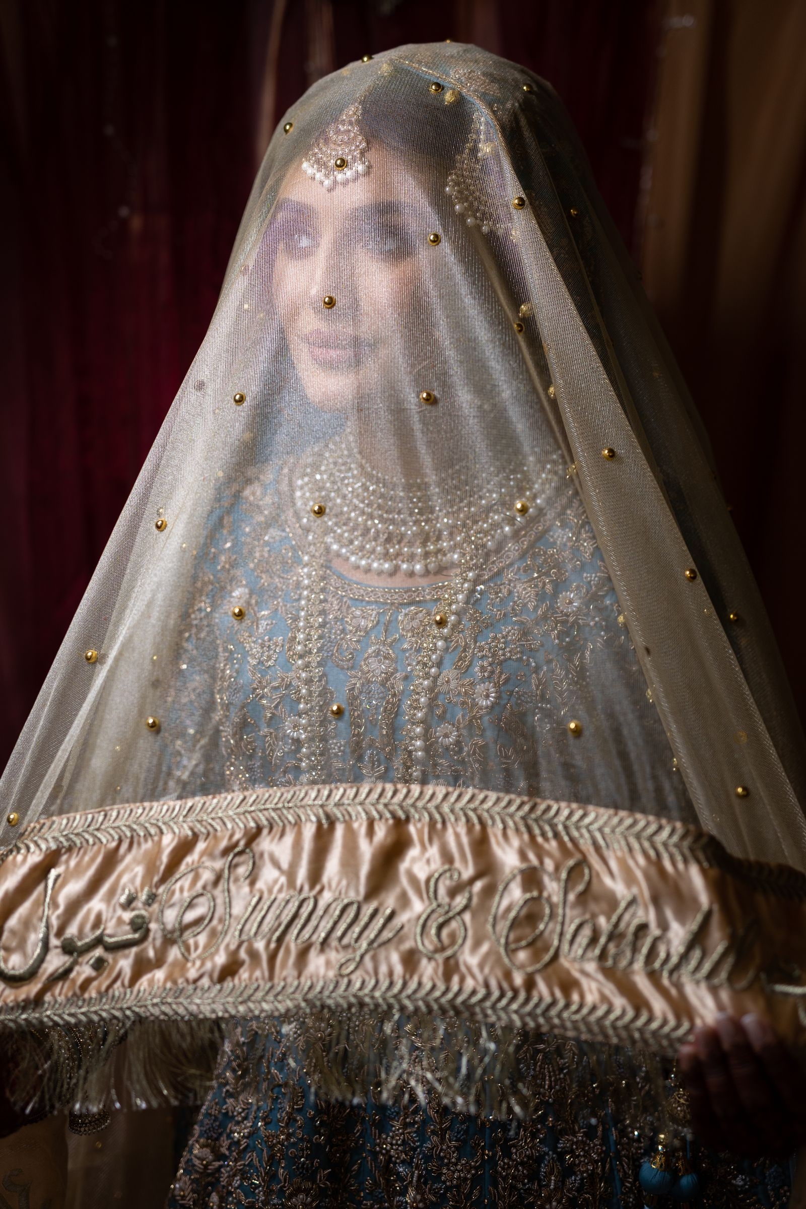 Bride beneath an embroidered veil held over her, her face just visible through it