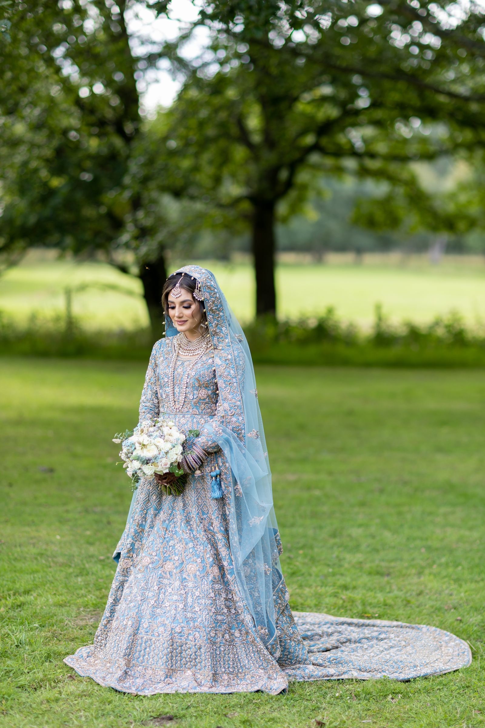 Bride standing on the lawn in a pale blue lehenga, her embroidered train spread behind her
