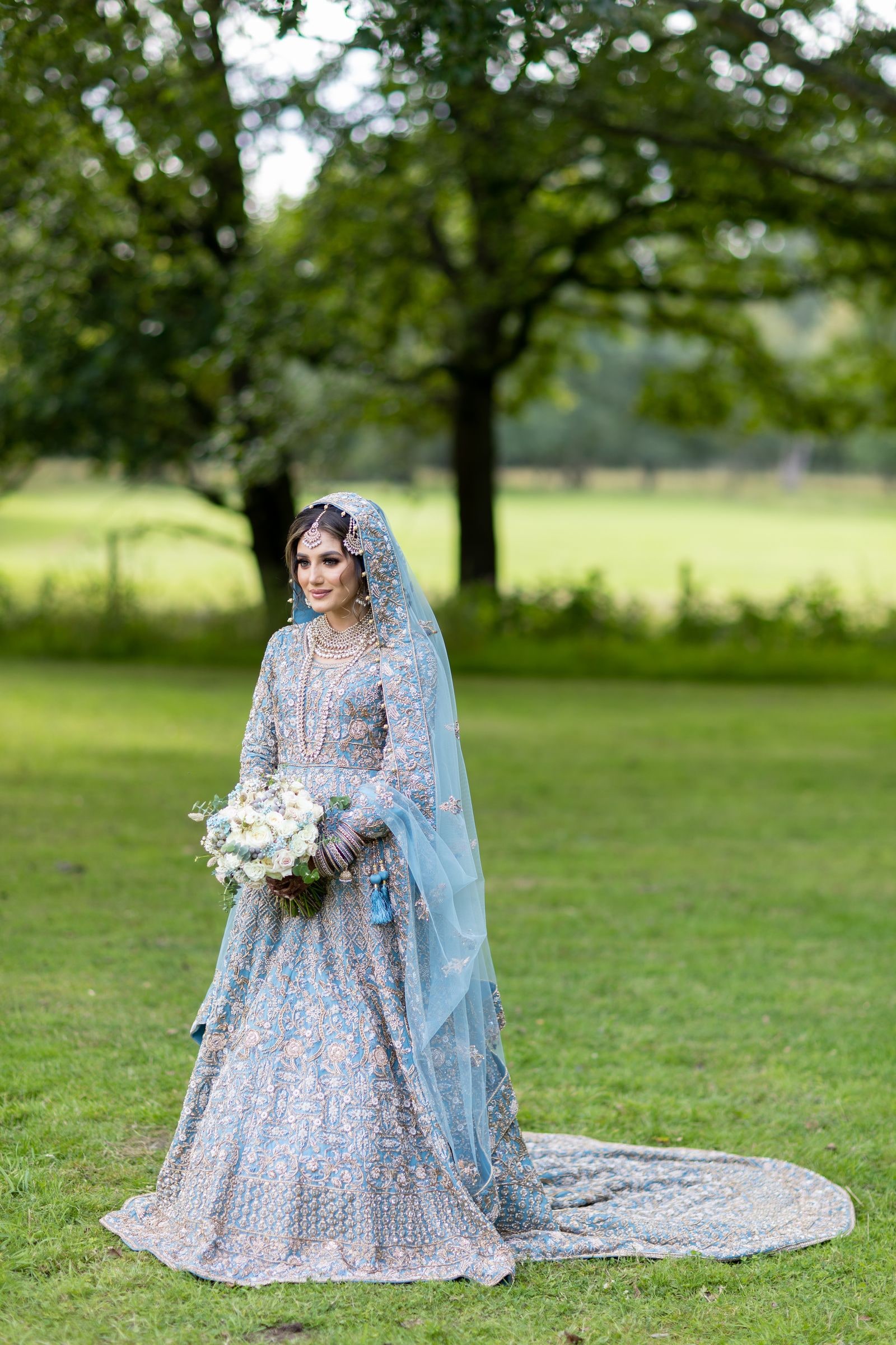 Bride under the trees holding a white bouquet, her train laid out on the grass