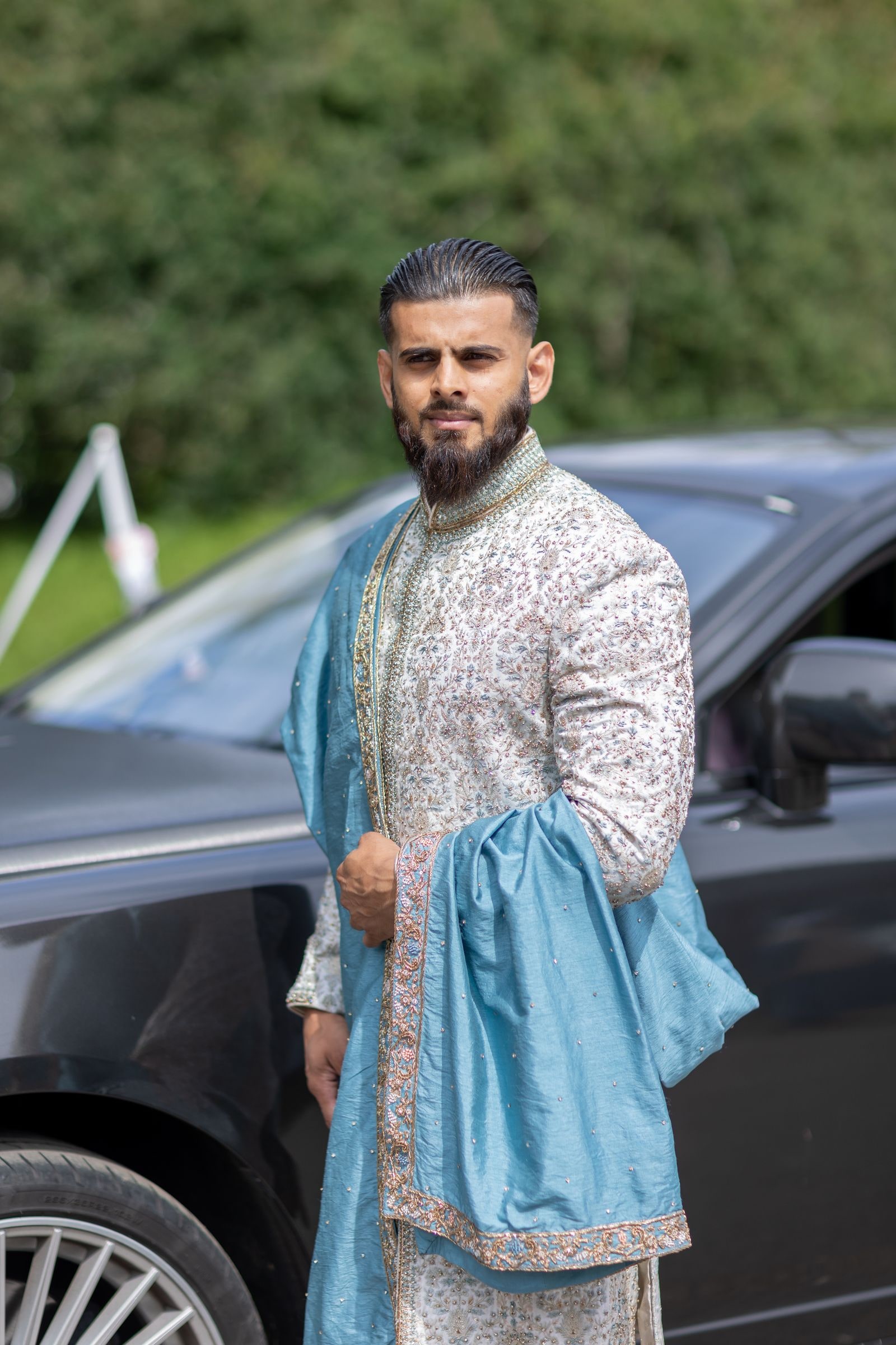 Groom in an ivory sherwani and blue shawl standing beside the car on arrival