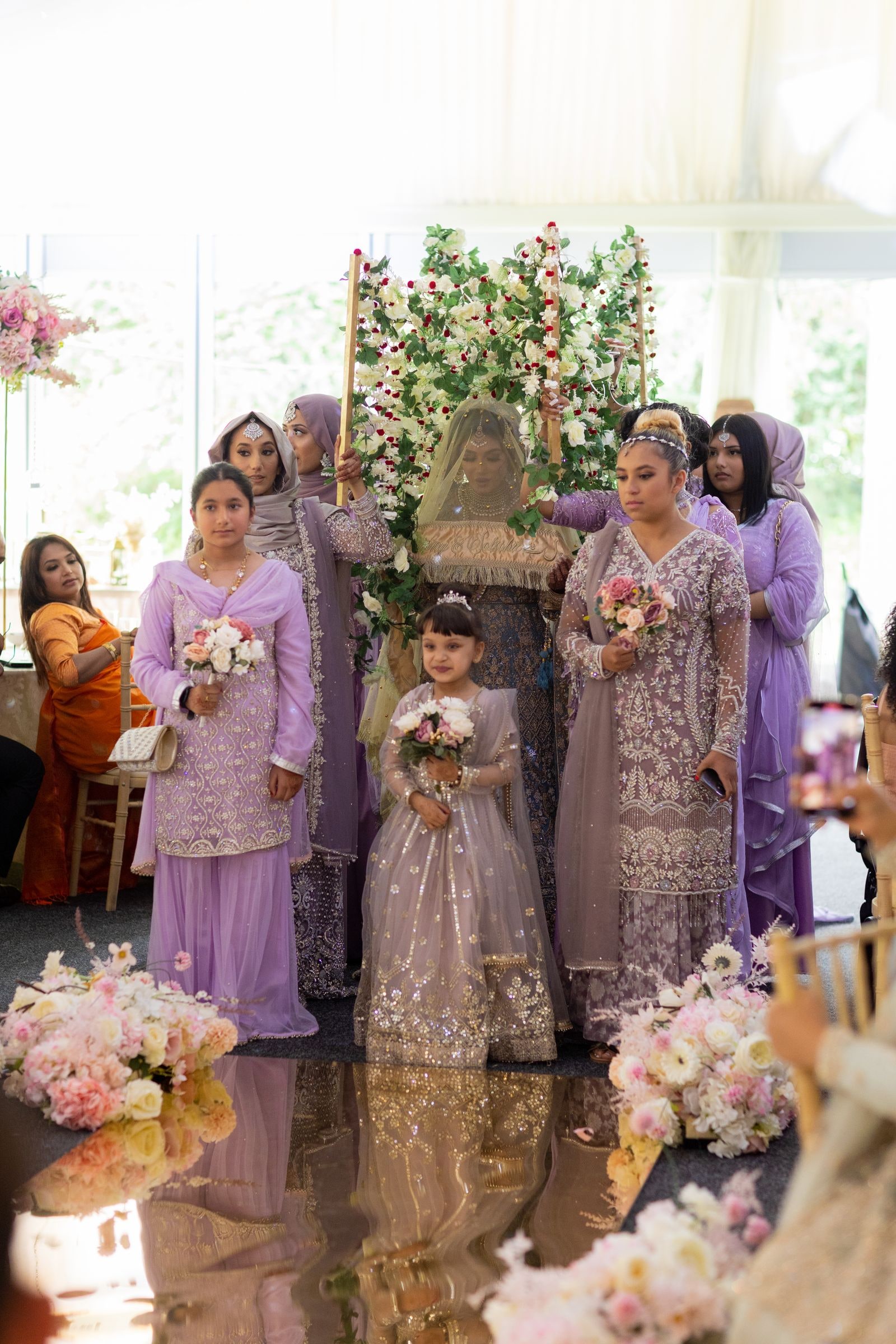 Bride veiled beneath the flowered canopy, her party in lilac around her