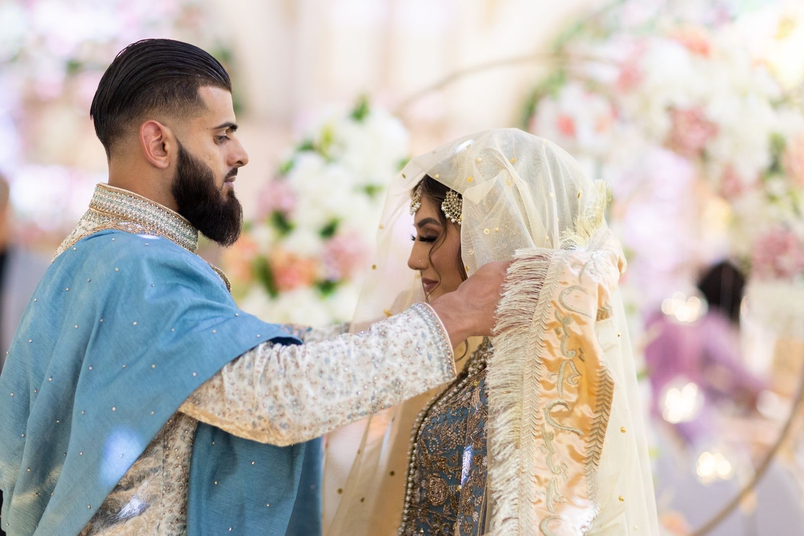 Groom lifting his bride's veil during the ceremony, flowers behind them