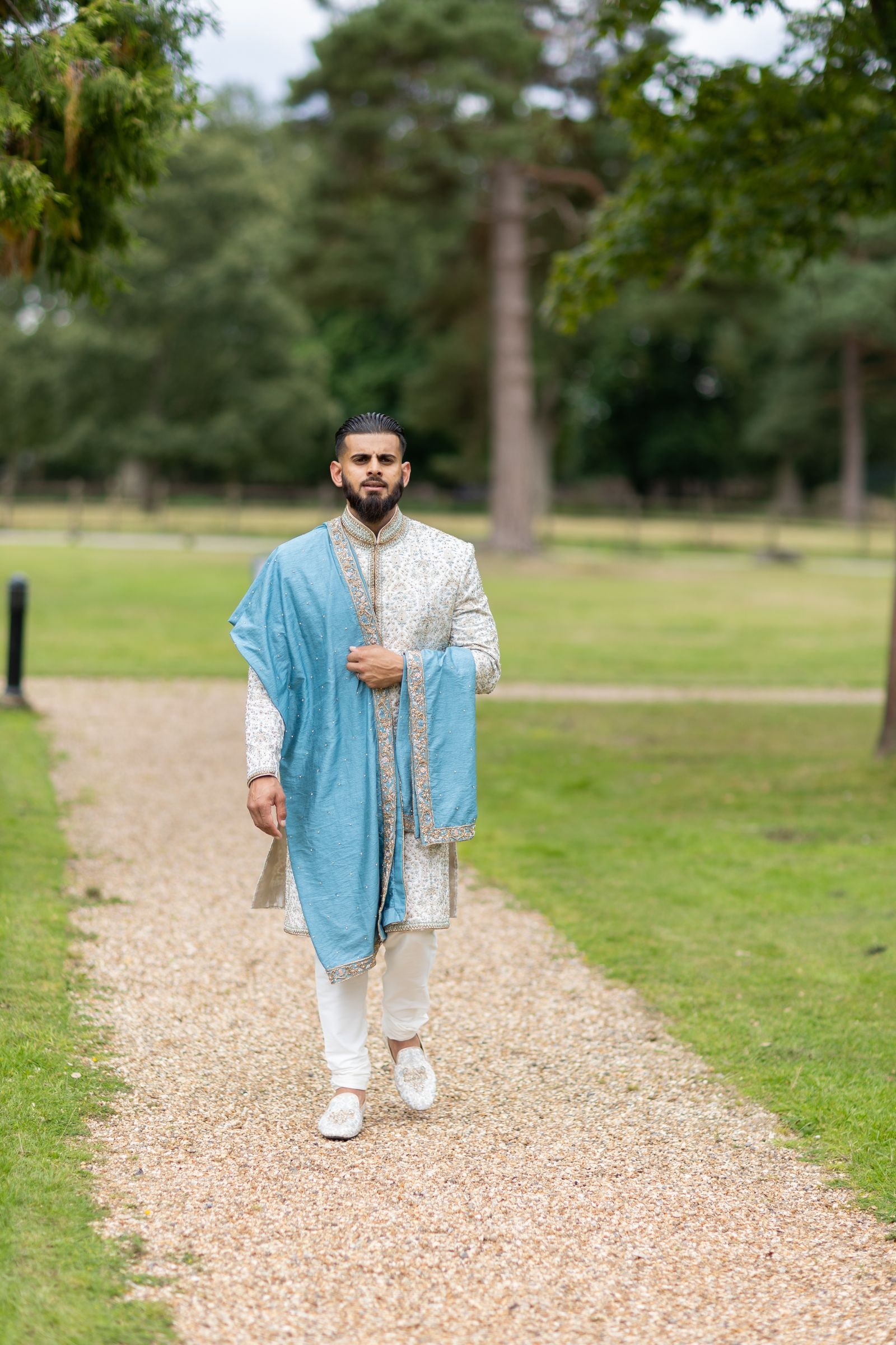 Groom walking up the gravel path alone in an ivory sherwani and blue shawl