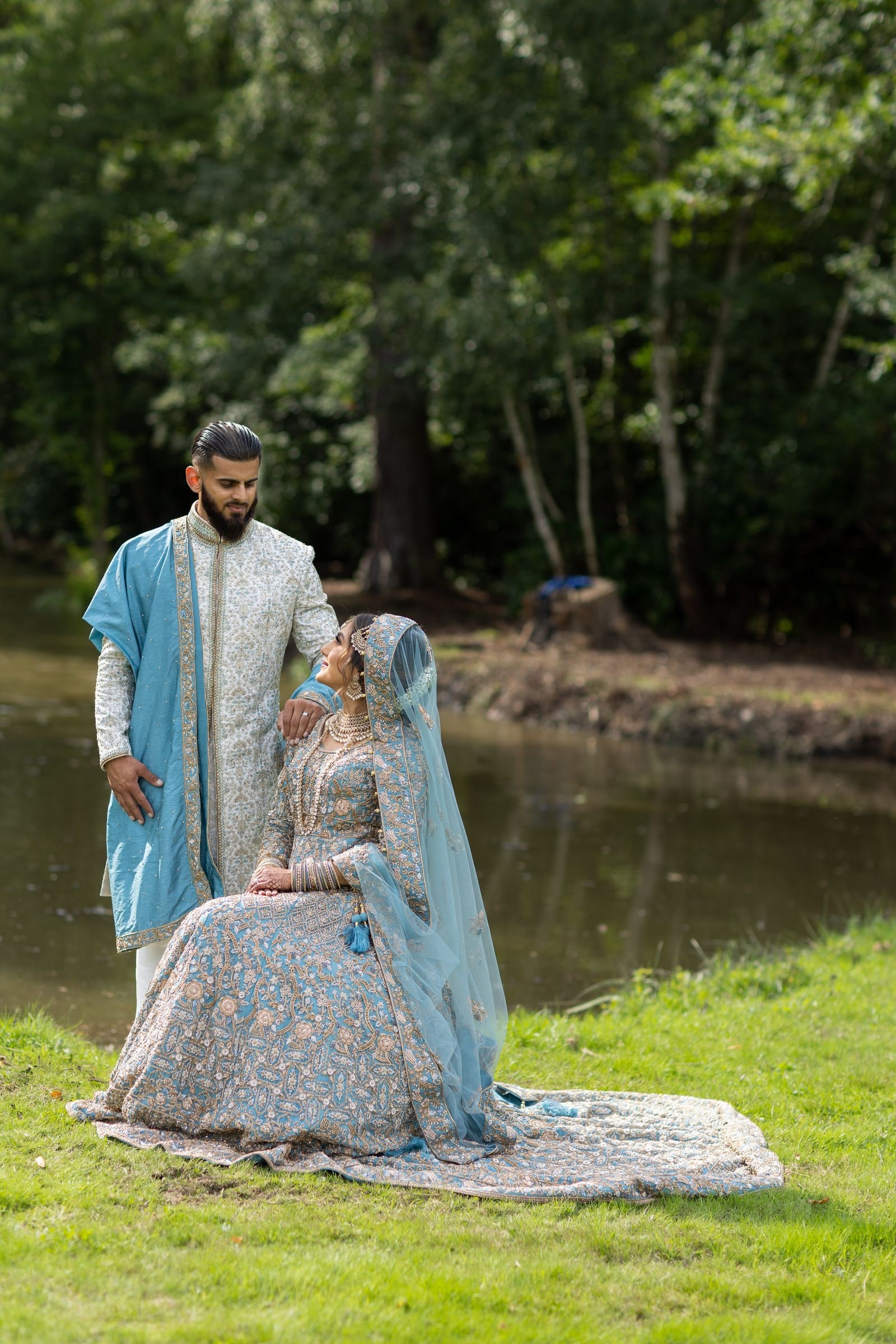 Bride seated on the grass by the lake, her groom standing over her