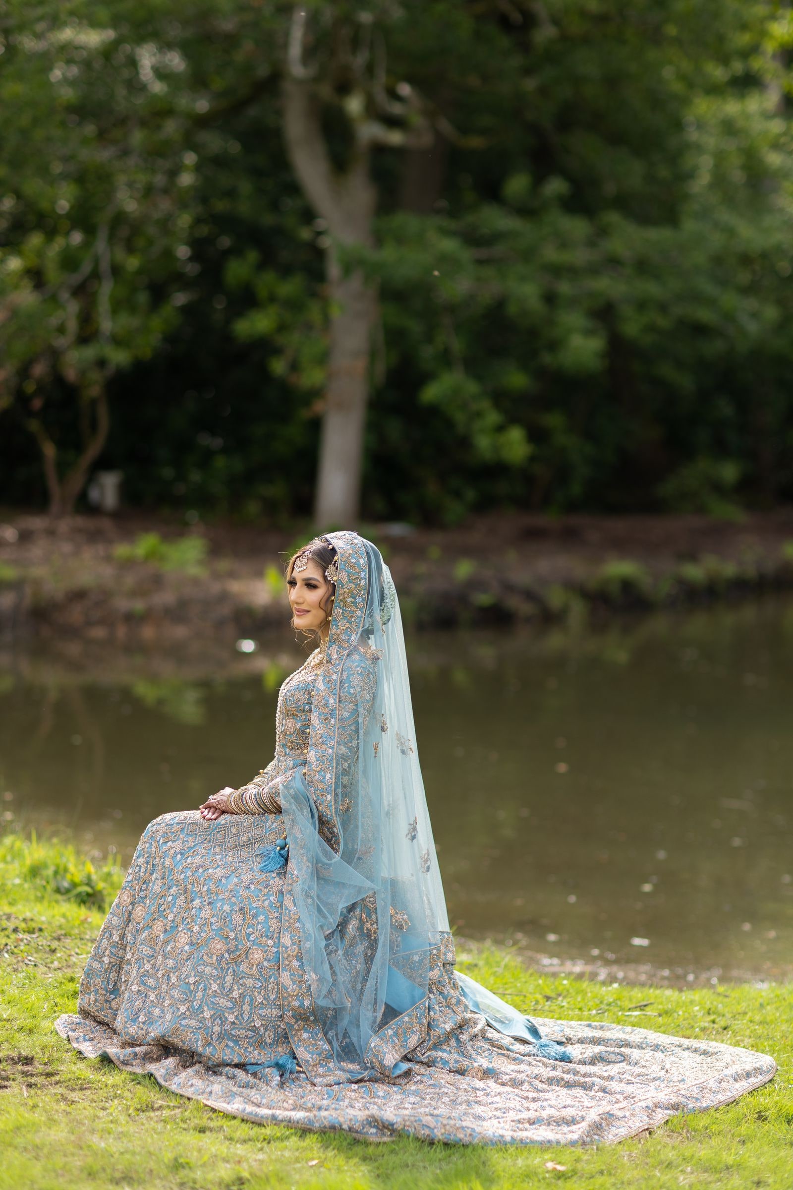 Bride seated on the bank in a pale blue lehenga, her train laid out on the grass