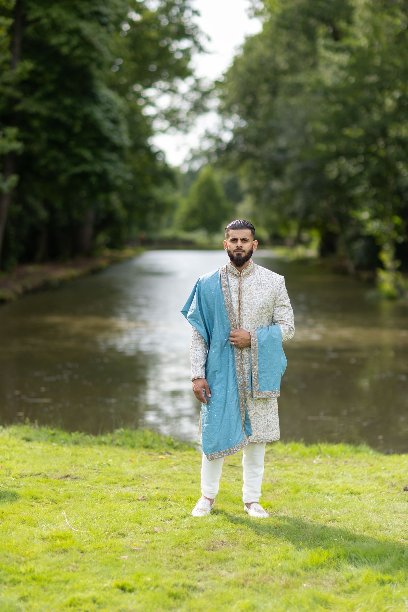 Groom standing alone on the bank in an ivory sherwani, the water behind him