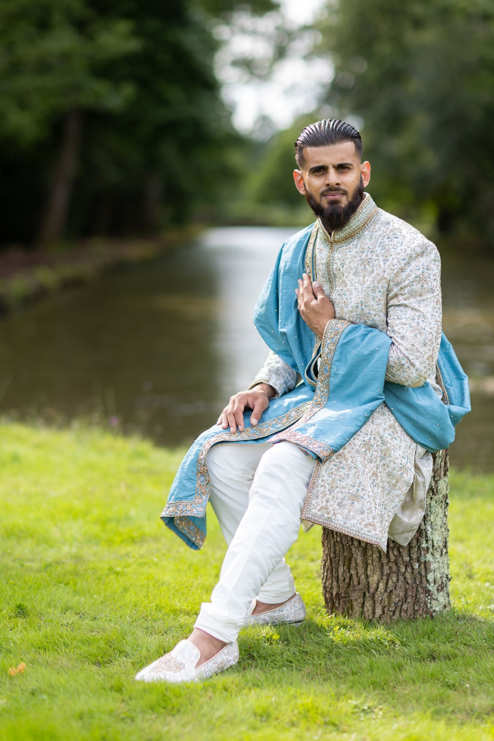 Groom seated on a tree stump by the water, his blue shawl over one shoulder