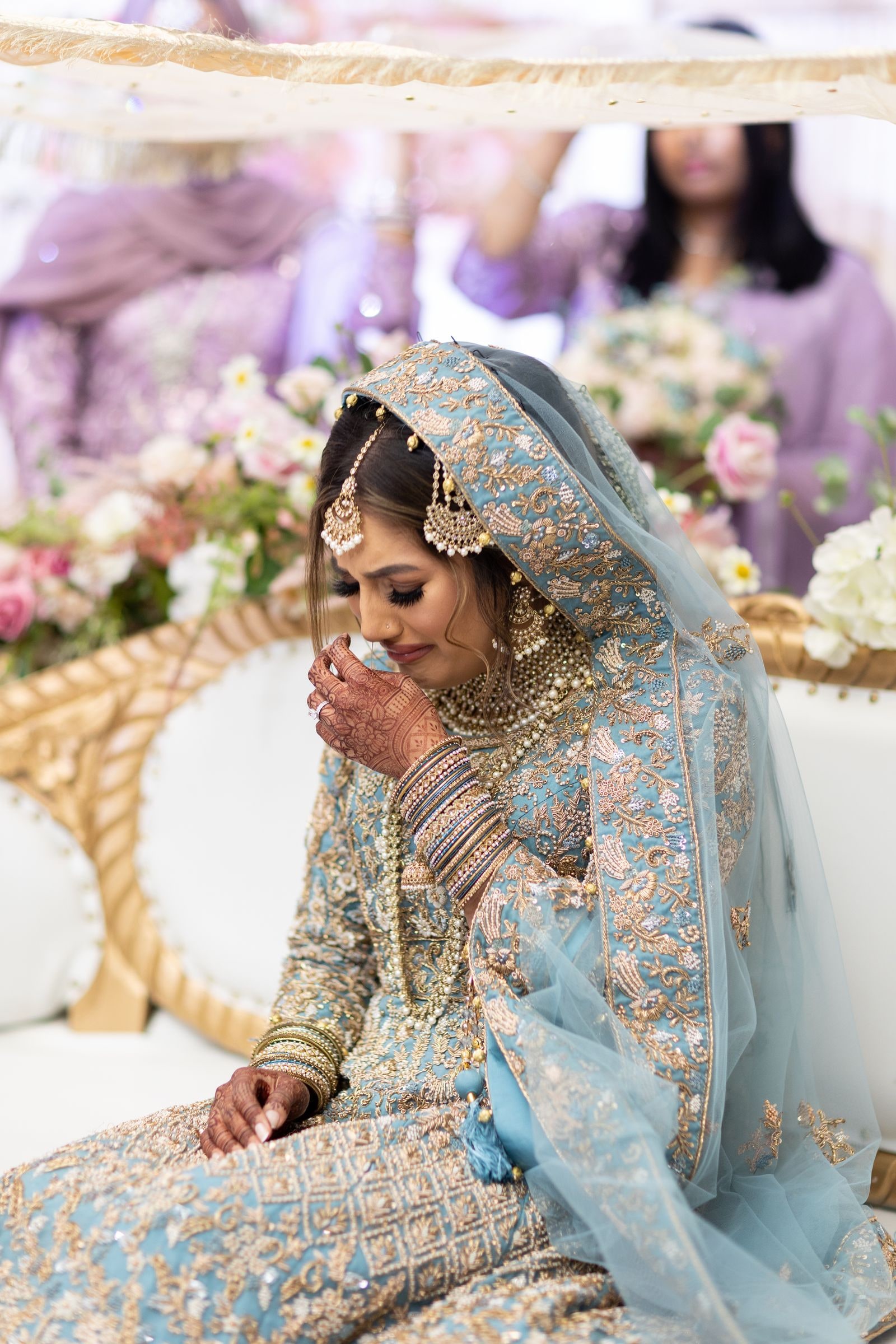 Bride seated on the stage wiping her eye, her party in lilac behind her