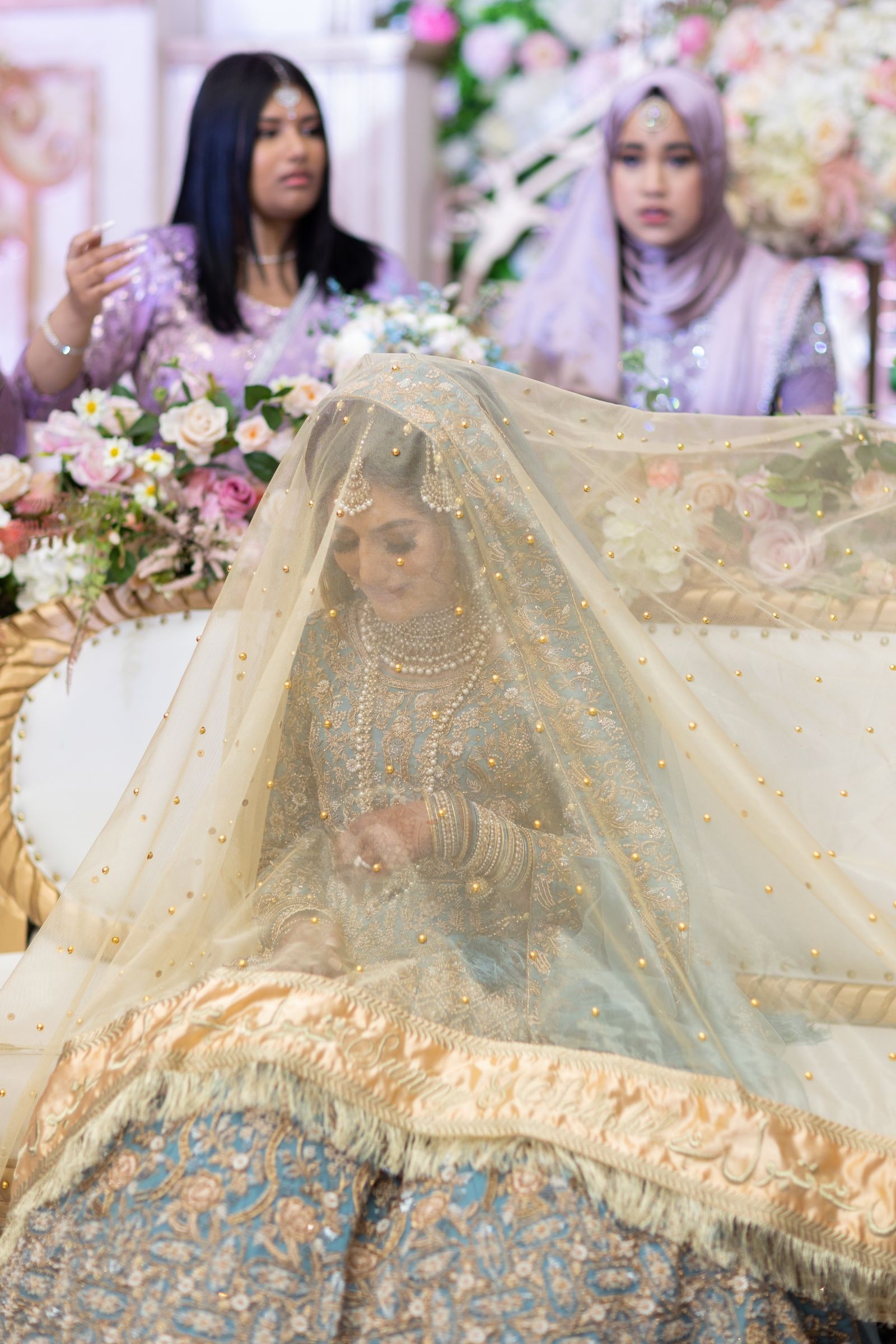 Bride seated under a gold veil, watched by two women in lilac