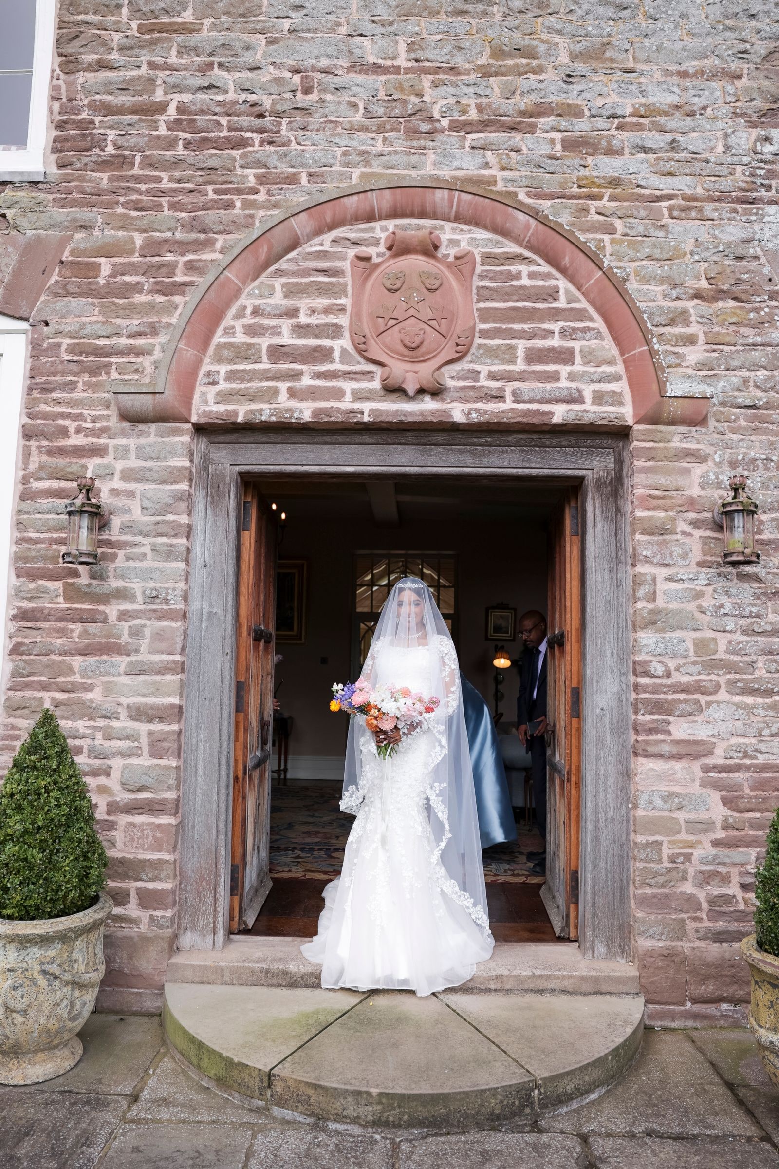 Bride standing veiled in the arched stone doorway of the house, bouquet in hand