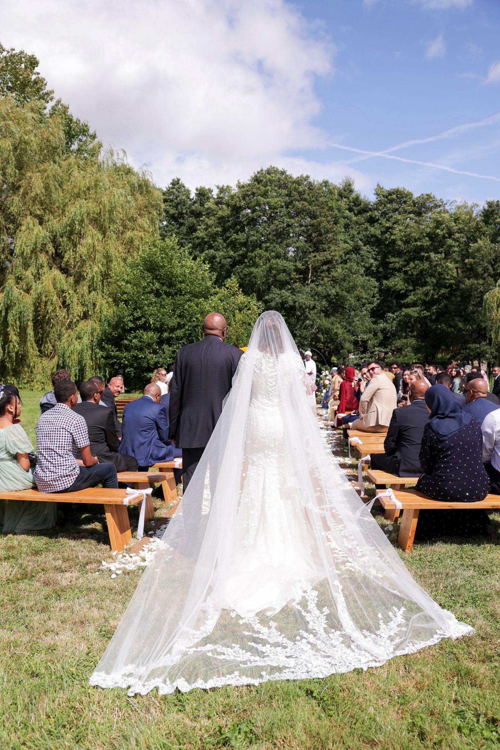 Bride walking the outdoor aisle from behind, her cathedral veil spread across the grass
