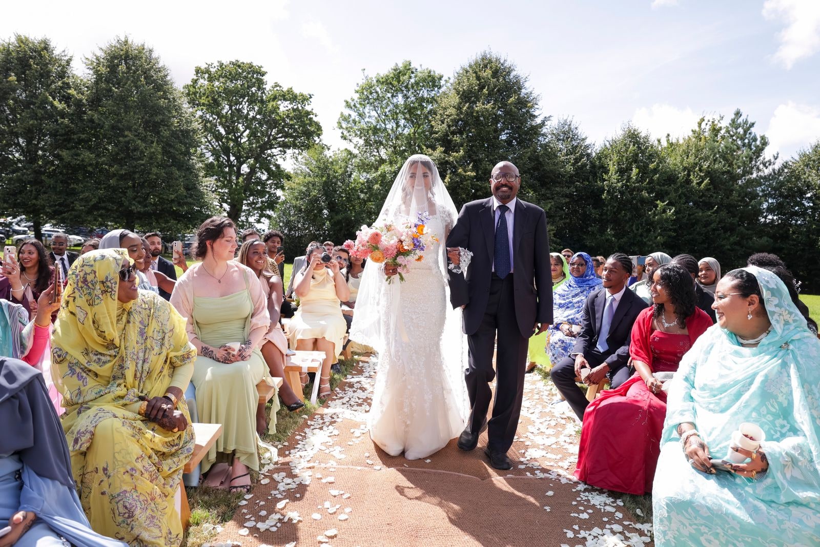 The bride walked down the outdoor aisle by her father, guests seated on benches