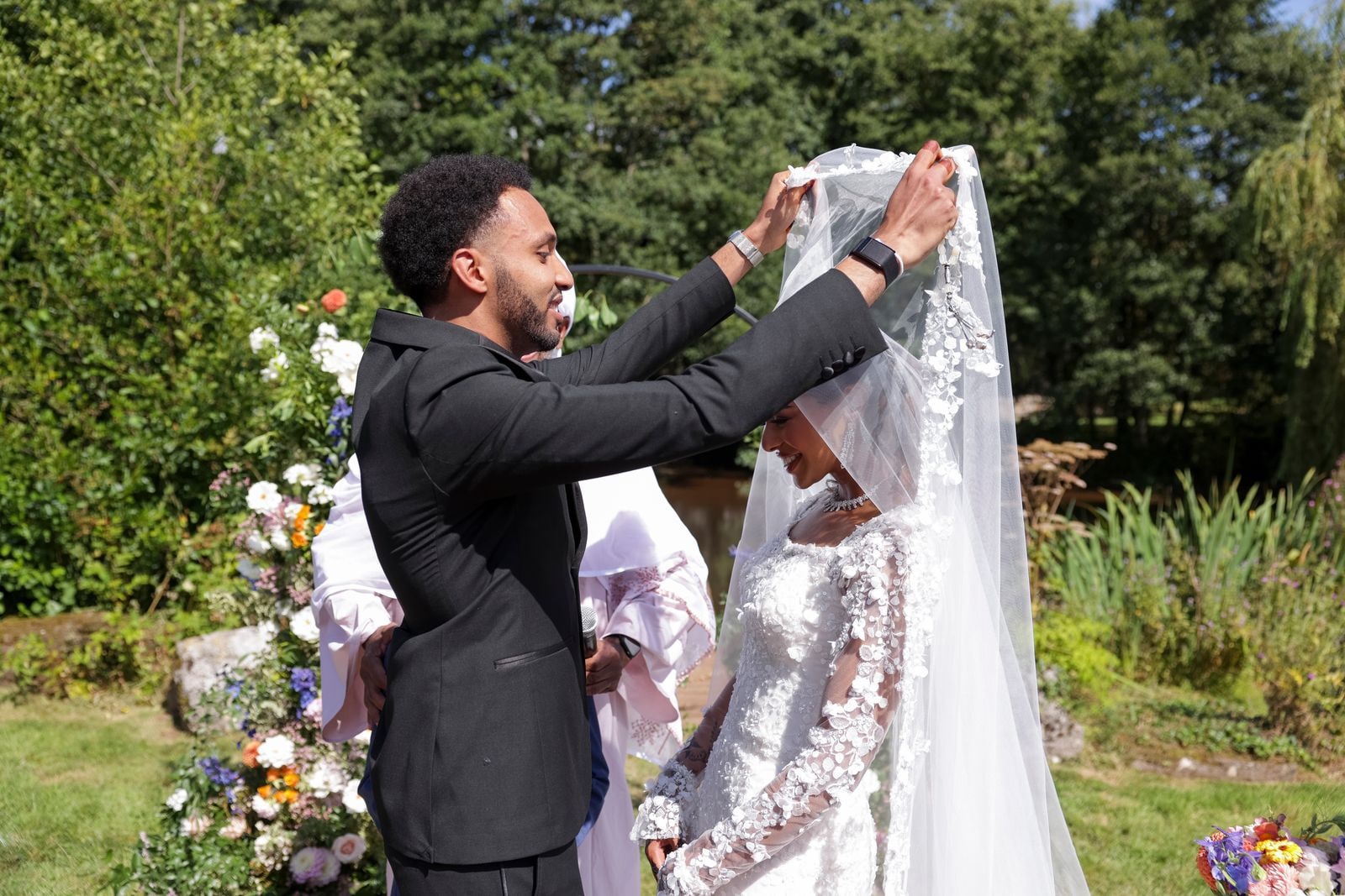 Groom lifting his bride's veil at the outdoor ceremony, flowers on the arch behind