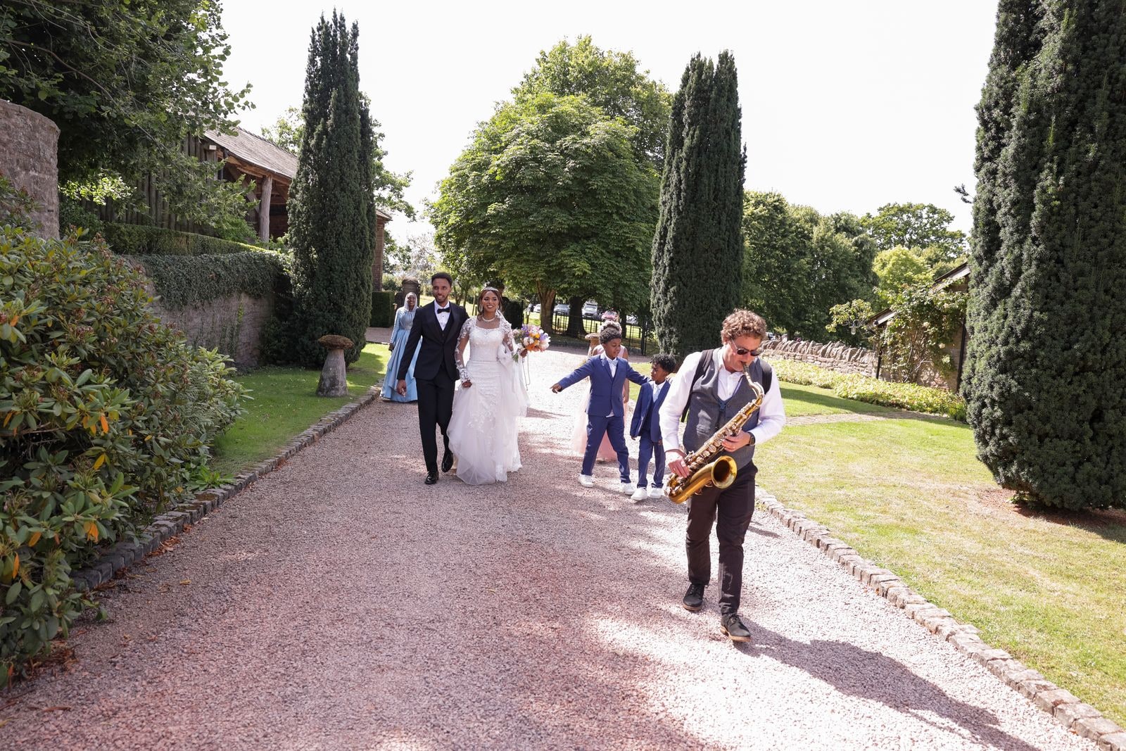 The couple walking the path behind a saxophonist and drummer between tall cypresses