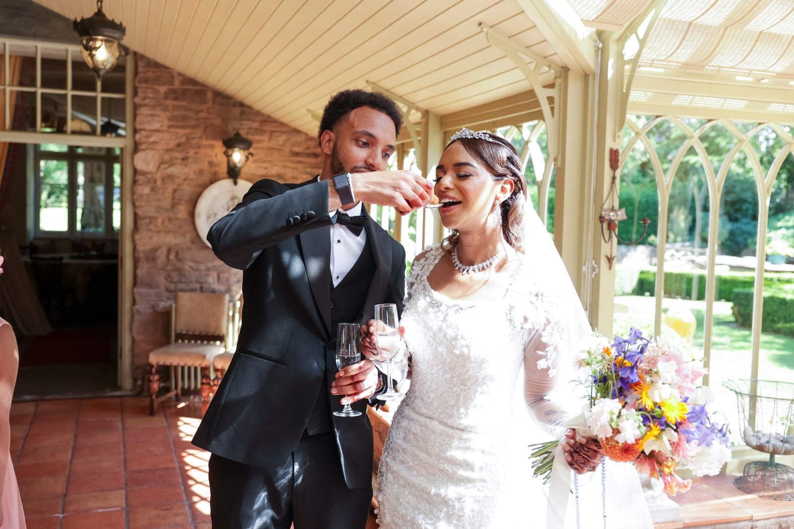 Bride and groom feeding each other in the conservatory, glasses in hand