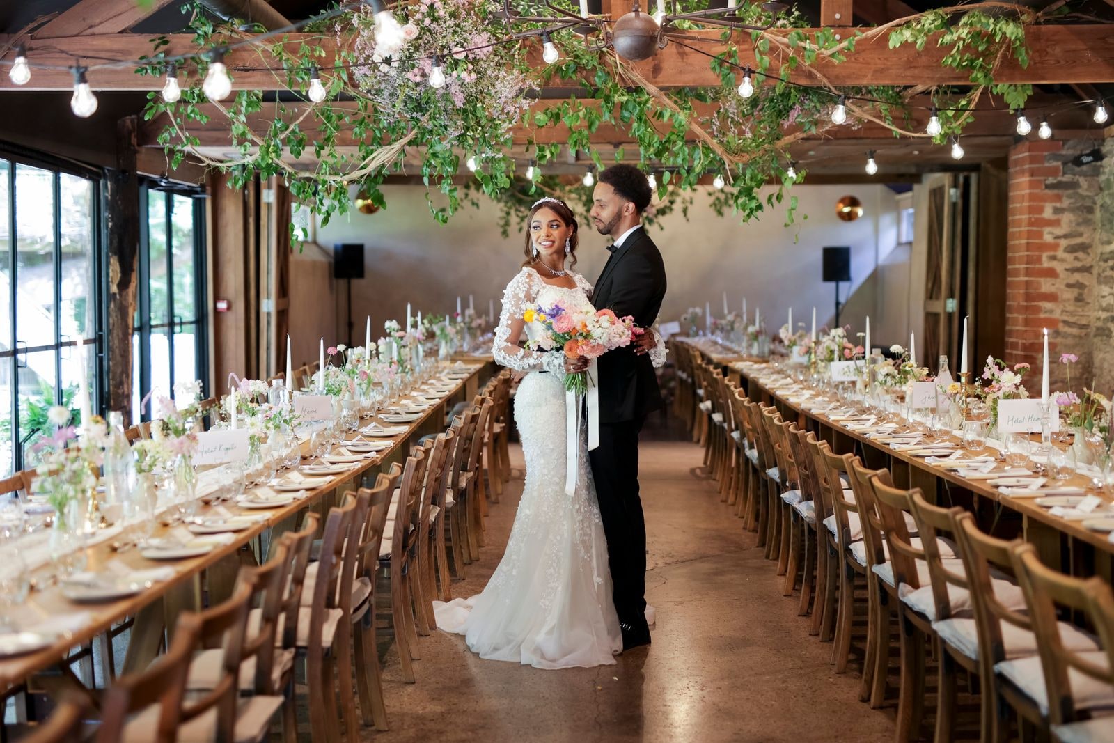 Bride and groom standing alone between the long tables in the barn